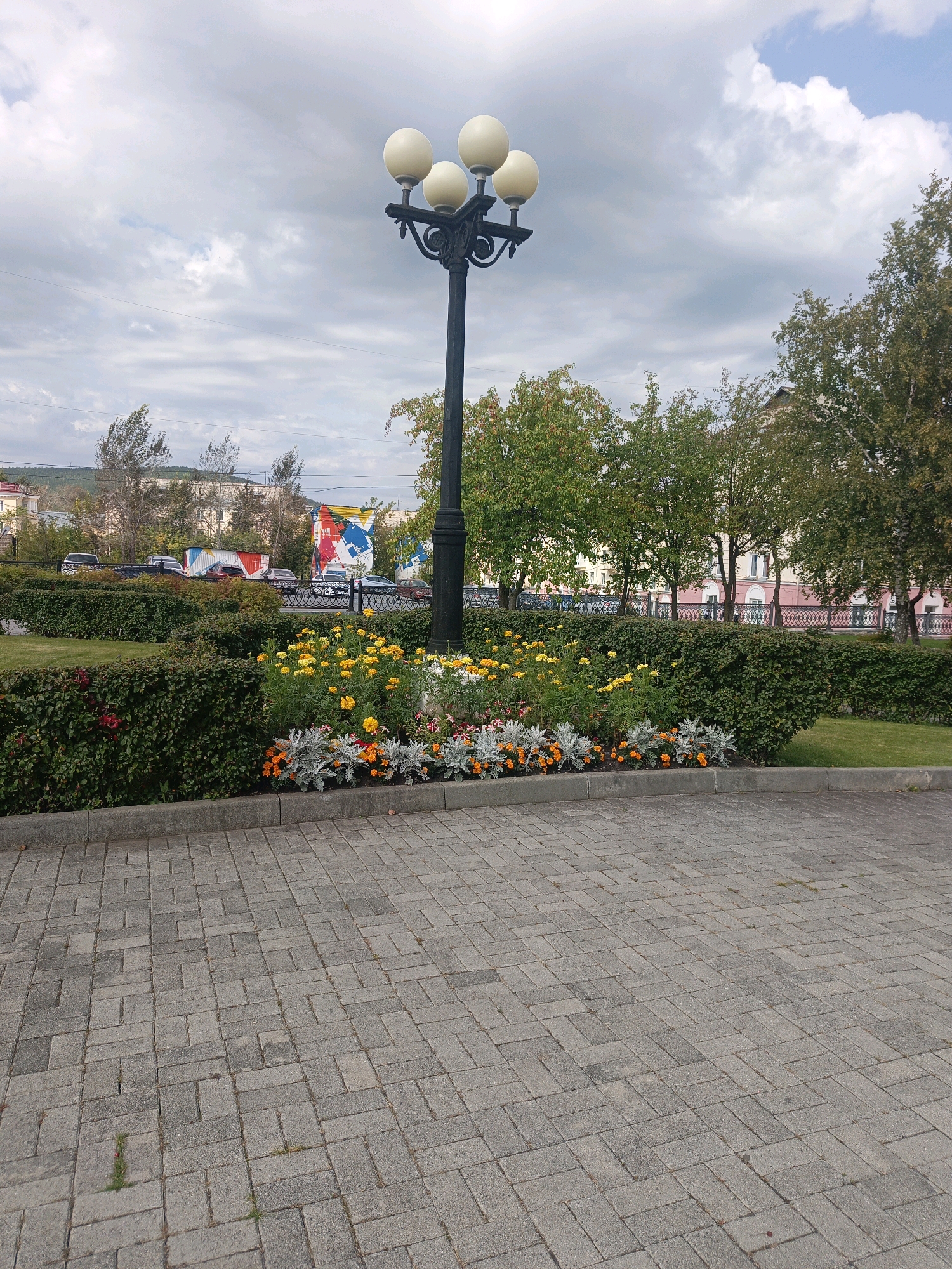 The image shows a street lamp with four light globes at the top, positioned in front of a flower bed filled with yellow and orange flowers, as well as silver-leafed plants. Behind the flower bed are trimmed green hedges, followed by a fence and a street with cars parked and driving. Beyond the street, there are buildings and trees. The sky above is cloudy with patches of blue. The foreground is paved with gray rectangular stones. The scene appears to be in the city of Satka, Russia, likely a public area or park given the landscaping and street furniture. There are no people visible in the image.
