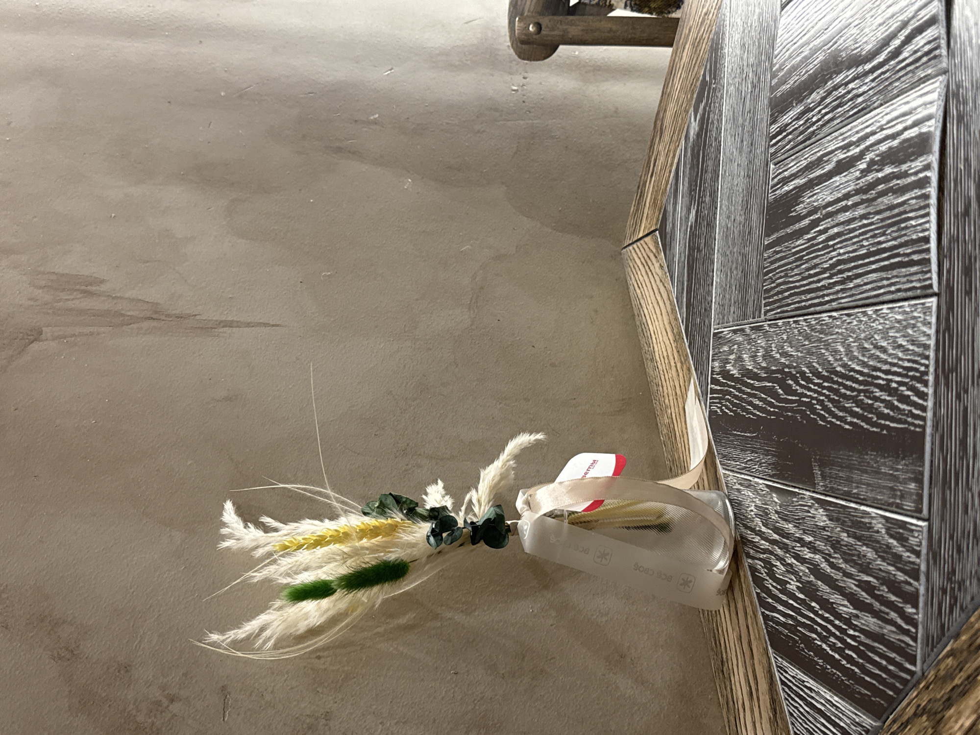 A close-up, eye-level, medium shot shows a dried floral arrangement in a clear glass vase, placed on a dark, wood-paneled table. The arrangement consists of white feathery plumes, green stalks, and yellow wheat-like stalks. A light-colored ribbon is tied around the vase, with a small tag attached. The background is a textured, beige wall with some dark, streaky marks. To the right of the frame, the corner of a wooden chair is visible. The scene appears to be indoors.