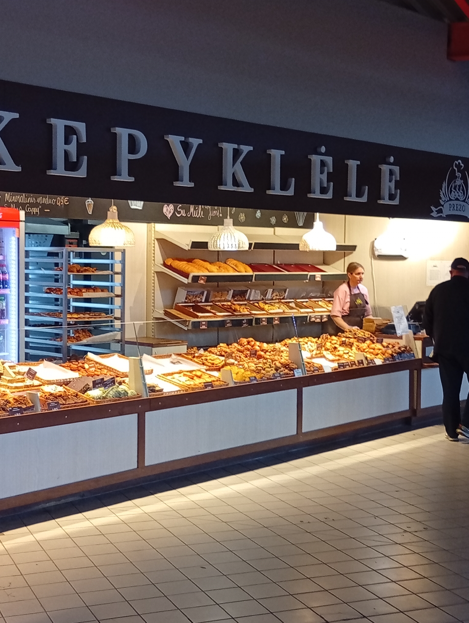 The image depicts the interior of a bakery, likely named "Kepykla" based on the prominent signage. The bakery is located in Šiauliai, Lithuania, as evidenced by the presence of the Lithuanian language on signs and products.
The scene shows a display of various baked goods, including breads, pastries, and cakes, arranged on shelves and in display cases. A female employee, wearing a pink shirt and a black apron, is visible behind the counter, interacting with customers or preparing items. A male customer, wearing a black jacket, is standing to the right of the frame, partially obscuring the view of the bakery's interior.
The bakery's interior is illuminated by overhead lights, casting shadows on the tiled floor. The overall impression is one of a bustling, well-stocked bakery.