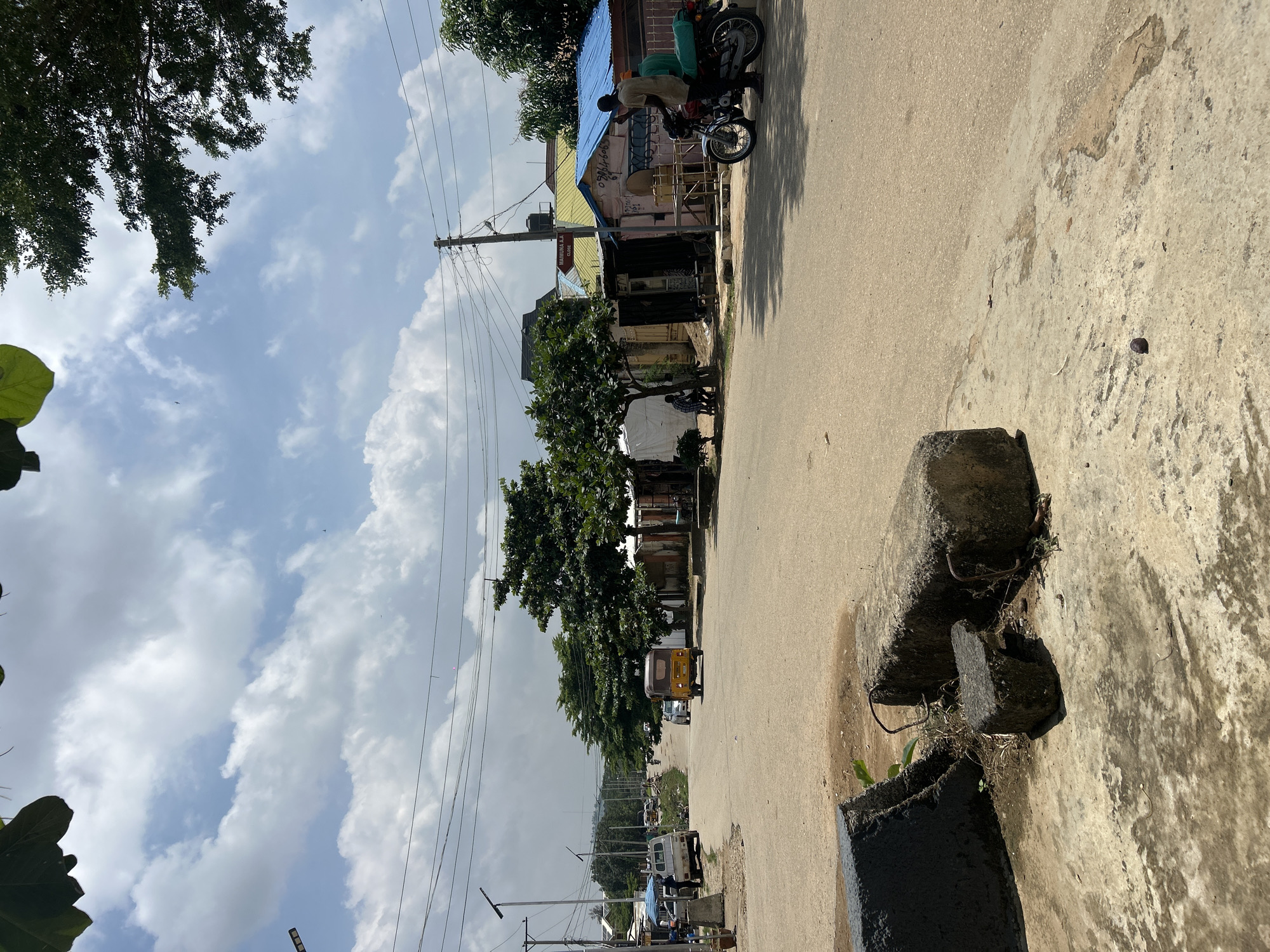 An eye-level, medium shot captures a street scene in Abuja, Nigeria. The paved road curves gently into the distance, lined with small shops and sparse vegetation. A yellow auto-rickshaw is visible in the middle of the road, moving away from the camera. On the right side of the road, two men are perched on a motorcycle. The sky above is a mix of blue and white clouds, with the upper branches of large trees framing the top of the image. In the foreground, on the left, are several large concrete blocks on the side of the road.