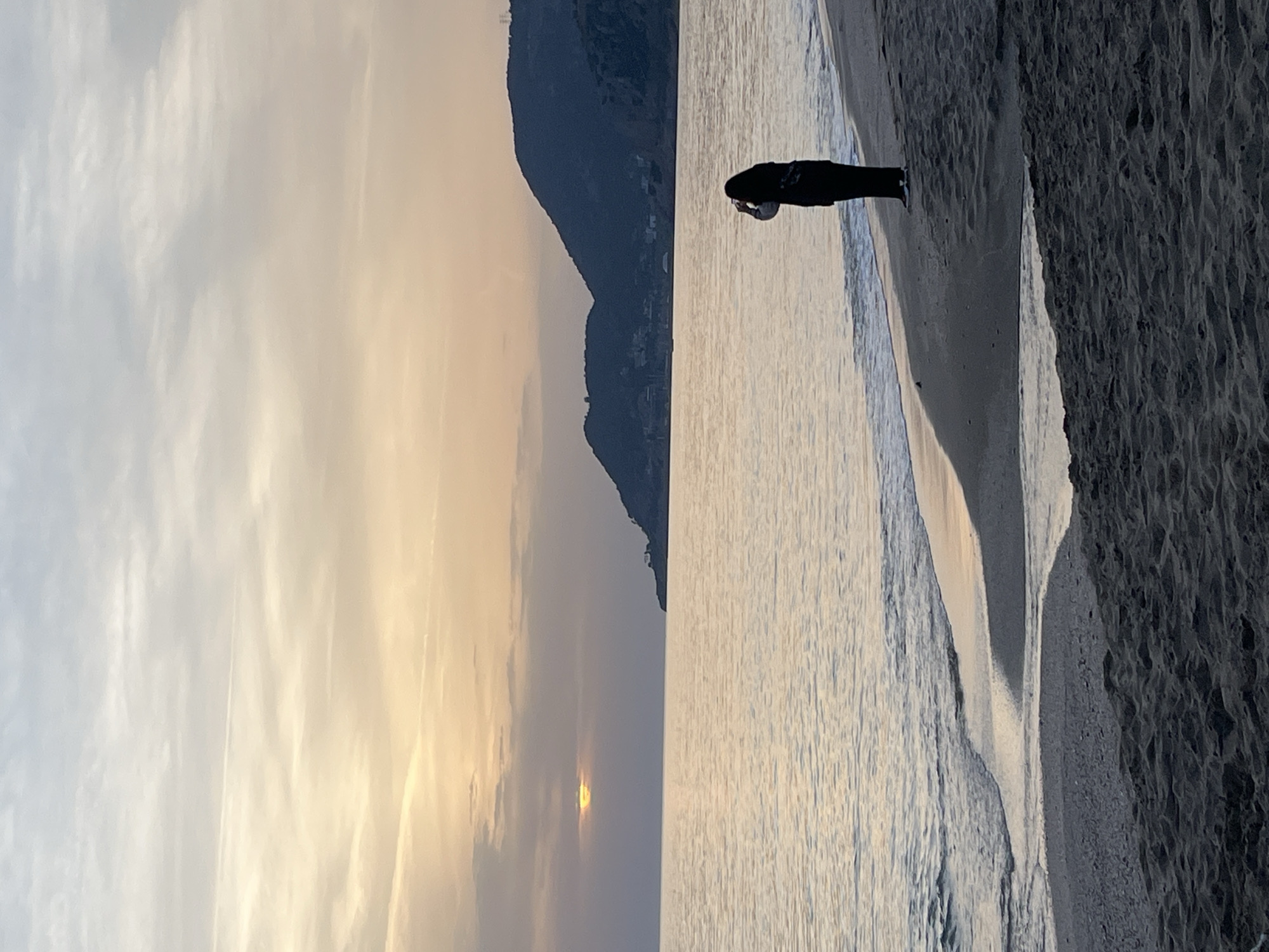 The image shows a beach scene, likely at sunset or sunrise. A person stands at the water's edge, facing the sea. The sky is bright with a gradient of colors, indicating the sun's position near the horizon. To the left, land formations are visible, suggesting the coastal terrain of Alanya, Turkey. The beach itself has dark sand, and the water appears calm. The person's silhouette is dark against the bright water.