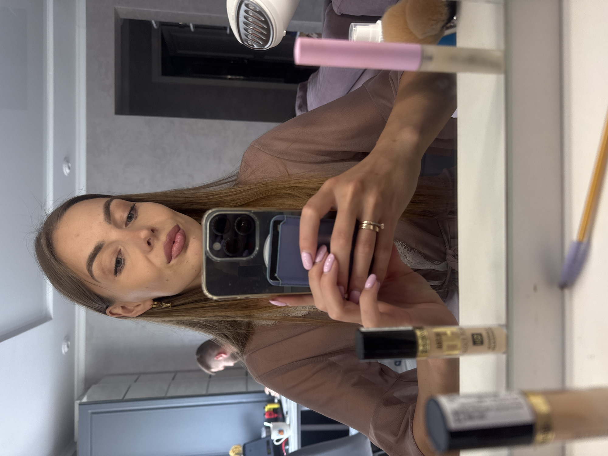A young woman with long brown hair is taking a selfie in a mirror. She is wearing a light brown, sheer robe with lace trim. She has on makeup, including lipstick and what appears to be nail polish. She is wearing two rings on her right hand, one of which is a thin gold band. In the foreground of the mirror reflection, there are several makeup products visible, including two tubes of concealer and a pink brush. In the background, a man with short brown hair is visible, looking away from the camera. The environment appears to be an indoor space, possibly a dressing room or bathroom, with grey walls and cabinetry. There is a white hairdryer on a shelf to the right. This scene is not located in Sochi, Russia; it is an indoor setting, likely a home or studio.