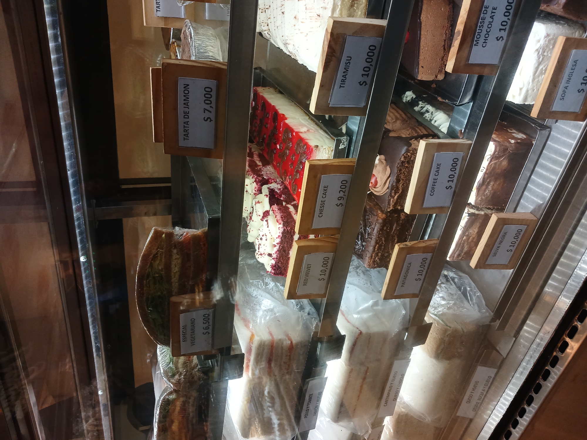 A refrigerated display case contains slices of various cakes and pastries. Each slice is labeled with a wooden tag displaying the type of cake and the price, denoted in pesos. The selections include "Especial Vegetariano" ($6,500), "Tarta de Jamon" ($7,000), "Chesse Cake" ($9,200), "Red Velvet" ($10,000), "Tiramisu" ($10,000), "Coffee Cake" ($10,000), "Mousse de Chocolate" ($10,000), "Patagonica" ($10,000), "Selva Negra" ($10,000), and "Sopa Inglesa" ($10,000). Sandwiches are also included in the case. Some of the sandwiches are labeled as "Pollo y Tomate/Jamon y Huevo" and "Cantimpalo y Queso/Queso y Choclo" and "Queso y Huevo/Crudo, Rucula y Queso". The location is likely a bakery or cafe in Buenos Aires, Argentina, based on the Spanish labels and the use of pesos.