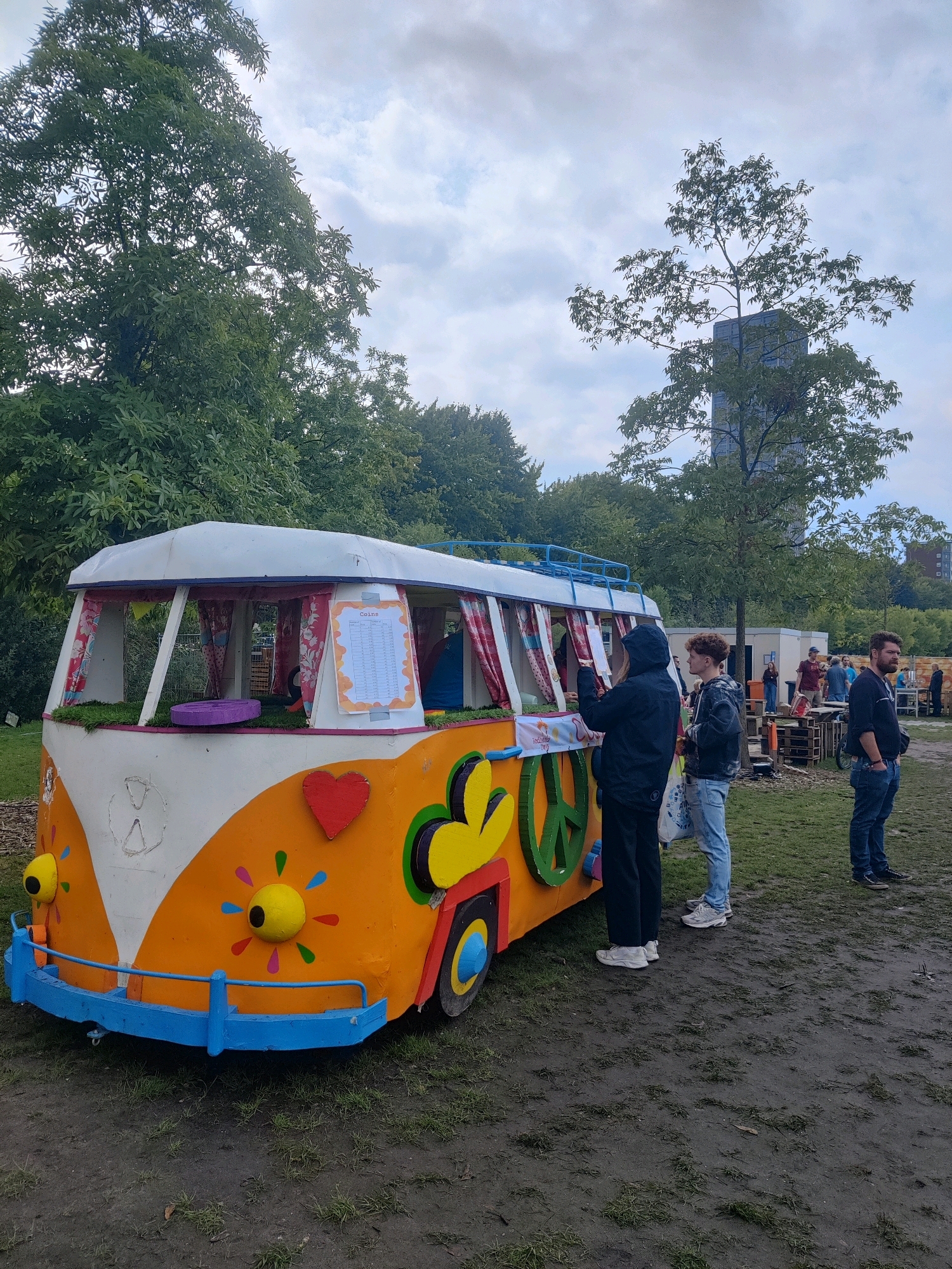 A colorful, retro-style van, painted orange with white and blue accents, is parked on muddy ground. The van is decorated with flowers, a heart, and a peace sign. Several people are standing around the van, some appearing to be interacting with it. In the background, there are trees, a tall building, and other structures that suggest an outdoor event or gathering. The sky is overcast with clouds. The van appears to be a food or drink stand, with a menu visible on its side. The scene suggests a casual, outdoor event, possibly a festival or market, taking place in an urban park or open area. The specific location is confirmed as Tilburg, Netherlands, at the Spoorzone area, near the former railway workshop.