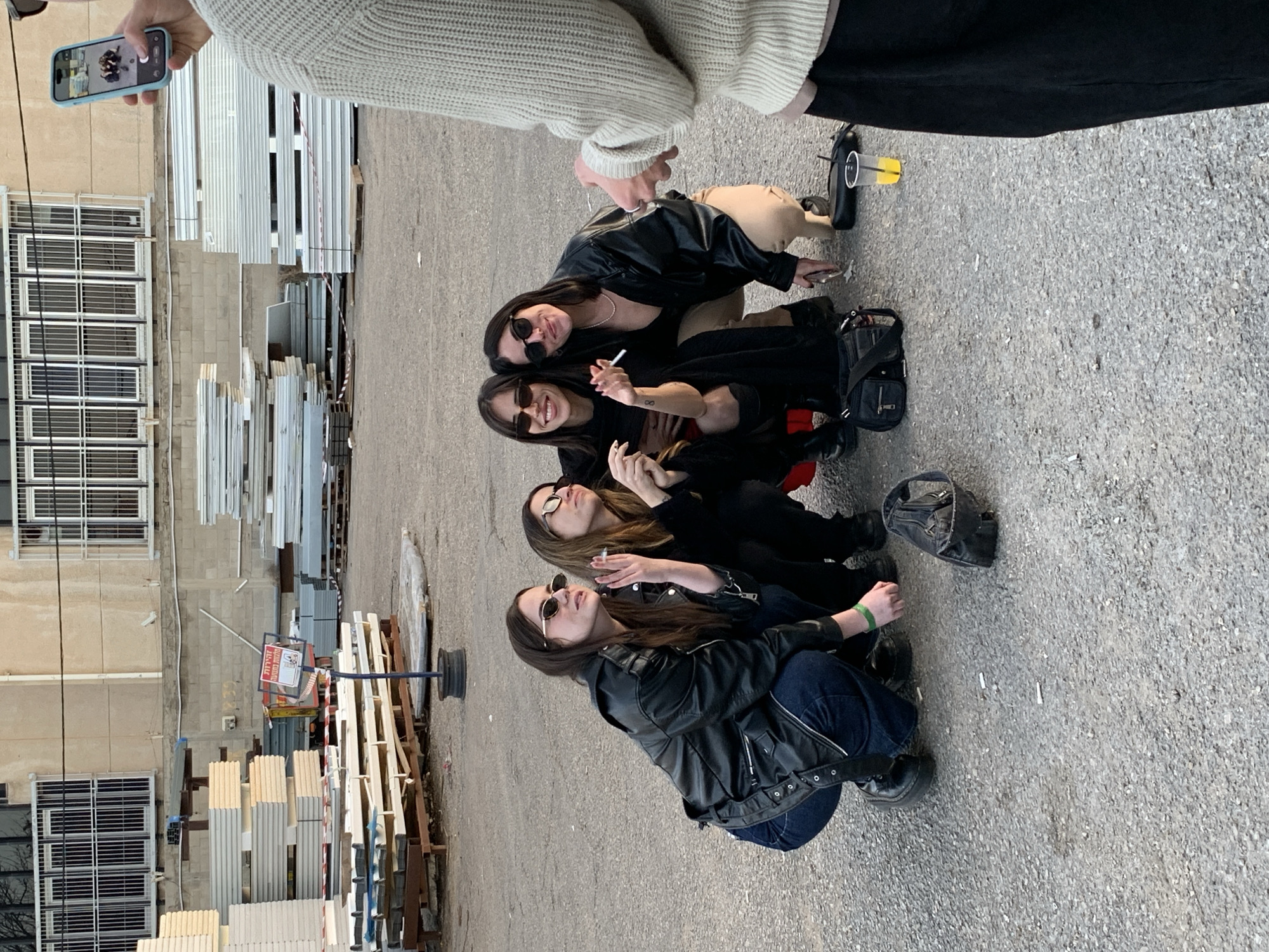 The scene is set in an industrial or commercial yard in Tel-Aviv, Israel. Four young adult women are crouched together on a grey asphalt surface, posing for a photograph. They are all wearing sunglasses, dark clothing, primarily black leather jackets and dark pants; one woman also wears beige pants. One woman on the far left wears a green wristband. The woman second from the left is holding a lit cigarette, and the woman next to her appears to be holding a cigarette as well. Two black bags, scattered cigarette butts, and a clear plastic cup with a yellow liquid and straw are on the ground near the women.
In the immediate right foreground, a person is partially visible, dressed in a light-colored knitted sweater and dark pants, holding a light blue smartphone horizontally to take the photograph. The phone's screen displays a live preview of the four women.
The background features stacks of various construction materials, including light-colored panels and grey metallic sheets, resting on wooden pallets. Behind these materials, a two-story building with a concrete facade and multiple windows secured by white metal grates is visible. A red and white sign, displaying Hebrew text that translates to "For Sale" and "Portable Buildings," is erected on a pole amidst the construction materials.