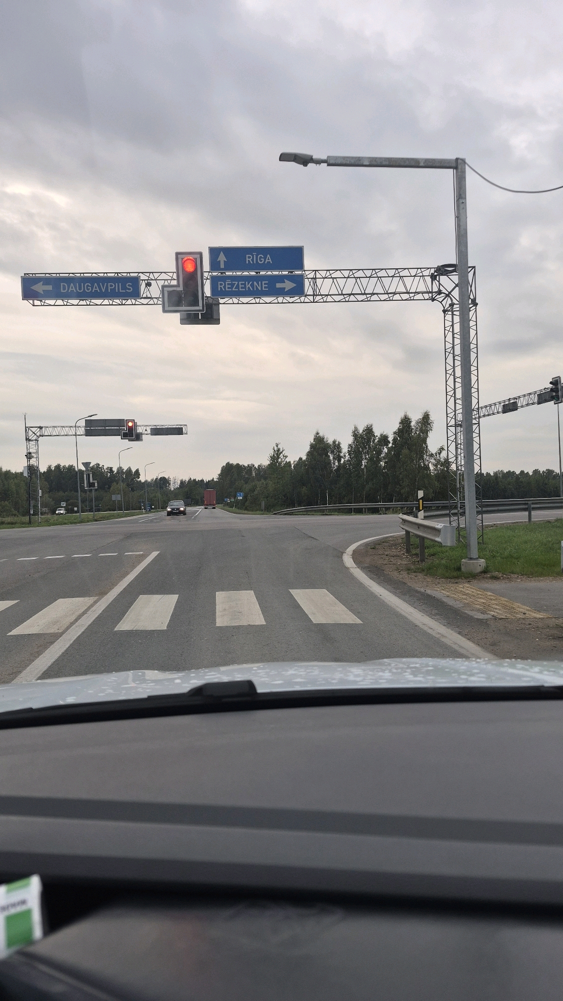 The image shows a view from inside a vehicle looking out at a road intersection in Lociki, Latvia, under an overcast sky. A traffic light is visible, displaying a red signal. Overhead signs indicate directions to Rīga, Daugavpils, and Rēzekne. The road has pedestrian crossings. In the distance, there are trees and a red truck. The foreground shows the dashboard of the vehicle.
