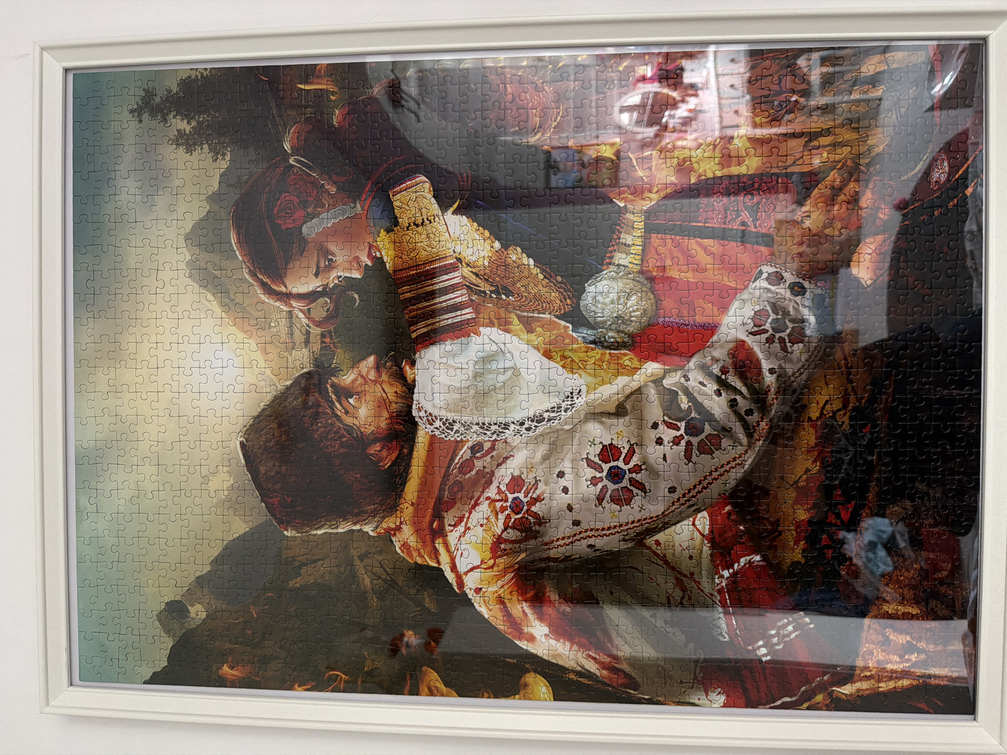The image depicts a completed jigsaw puzzle of an artistic portrayal of a couple, framed in white. The woman has red hair with a rose accessory, and the man has dark hair. They are dressed in richly decorated clothing with gold and red accents, positioned closely together. The background shows a landscape with trees and foliage. The reflection visible on the puzzle's surface suggests the image was taken indoors in a well-lit environment. There is no indication that this image is taken in Sofia, Bulgaria.