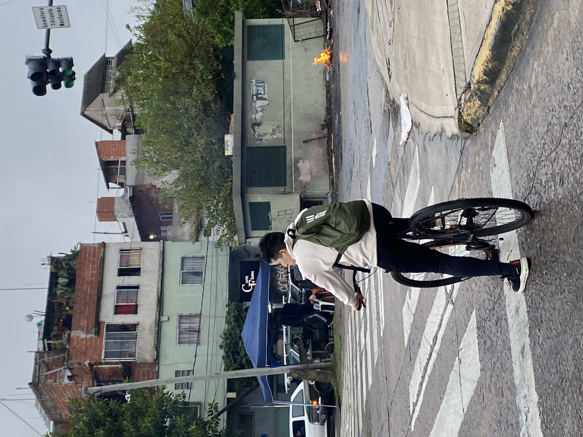 An urban street scene in Buenos Aires, Argentina, depicts a person riding a bicycle on a wet pavement, possibly after rain. The cyclist, dressed in a light-colored hoodie, dark pants, and a green backpack with an Adidas logo, is positioned on a zebra crossing. Their gaze is directed forward.
To the left, various buildings line the street, some appearing residential with visible windows and balconies, and some with exposed brickwork, contributing to a dense urban environment. Overhead, a traffic light displays a green signal and is accompanied by a sign that reads "SEMAFORO TITILANTE NOCTURNO" (Flashing Traffic Light Nocturnal).
In the mid-ground, a small, bright orange fire burns next to a weathered light-green building with dark green window shutters. The pavement around the fire is also wet. Further down the street, a blue tent structure is visible, possibly belonging to a market stall or temporary setup. Another building features a sign that reads "CORE Llama." The overall atmosphere is overcast, indicating a daytime shot without direct sunlight. The buildings show signs of wear and tear.