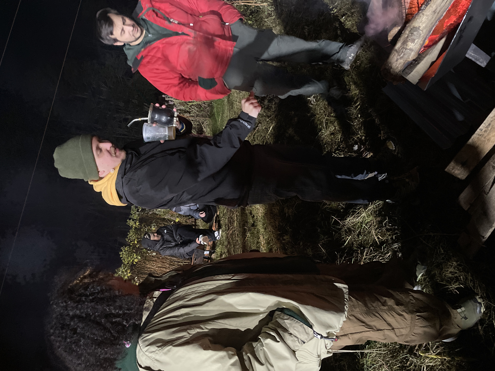 A group of people are gathered outdoors at night. Two men are lying down near a fire; one is wearing a red jacket, and the other is wearing a green hat and a black jacket over a yellow hoodie, and holding a drinking container with a straw. A partially visible individual in a brown jacket stands in the foreground. Another person is partially visible lying down in the background, wearing a dark jacket and white shoes. The location appears to be a grassy area, potentially in a rural or semi-rural environment. There is no information in the image to conclude this is in Ushuaia, Argentina.
