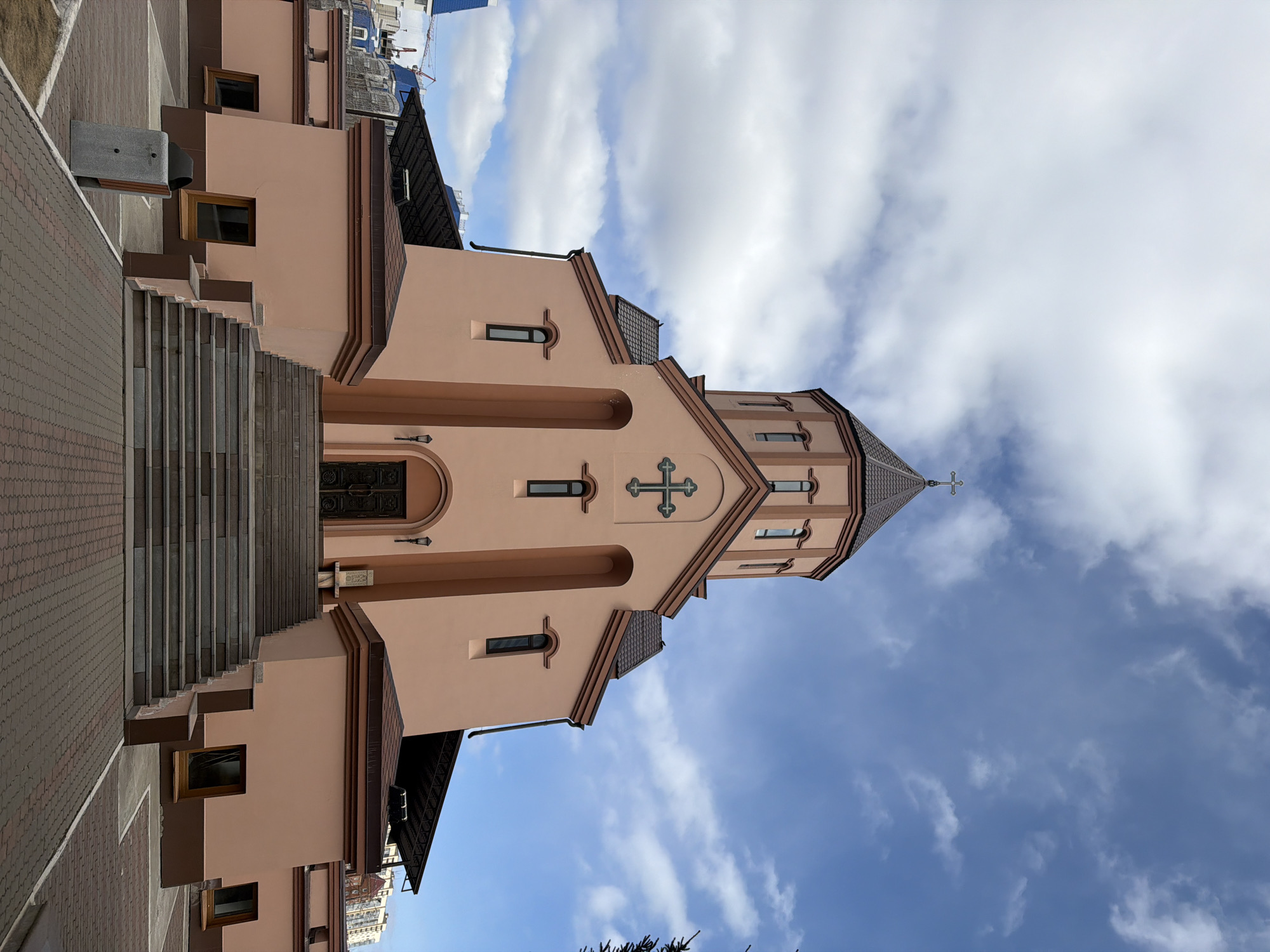 A medium-angle, eye-level shot captures a church building under a partly cloudy sky, likely in Krasnoyarsk, Russia. The church is predominantly painted a soft peach or pink hue, accented with brown trim along its rooflines and architectural features. Its main entrance is approached by a wide set of grey stairs on the left side of the frame, leading up to an ornate dark door.
Above the entrance, a decorative cross is prominently displayed on the facade. The building features several rectangular windows with dark frames and arched tops on its upper levels. The roof is multi-layered, culminating in an octagonal steeple topped with a metallic cross that reaches towards the sky. The architectural style suggests an Orthodox Christian design.
The ground around the church and stairs is paved with brown interlocking bricks. To the left of the stairs, a concrete bench or planter is visible. The sky is bright blue with scattered white, fluffy clouds, indicating daytime. No individuals or specific activities are visible, presenting a serene and static scene. No text is visible in the image.