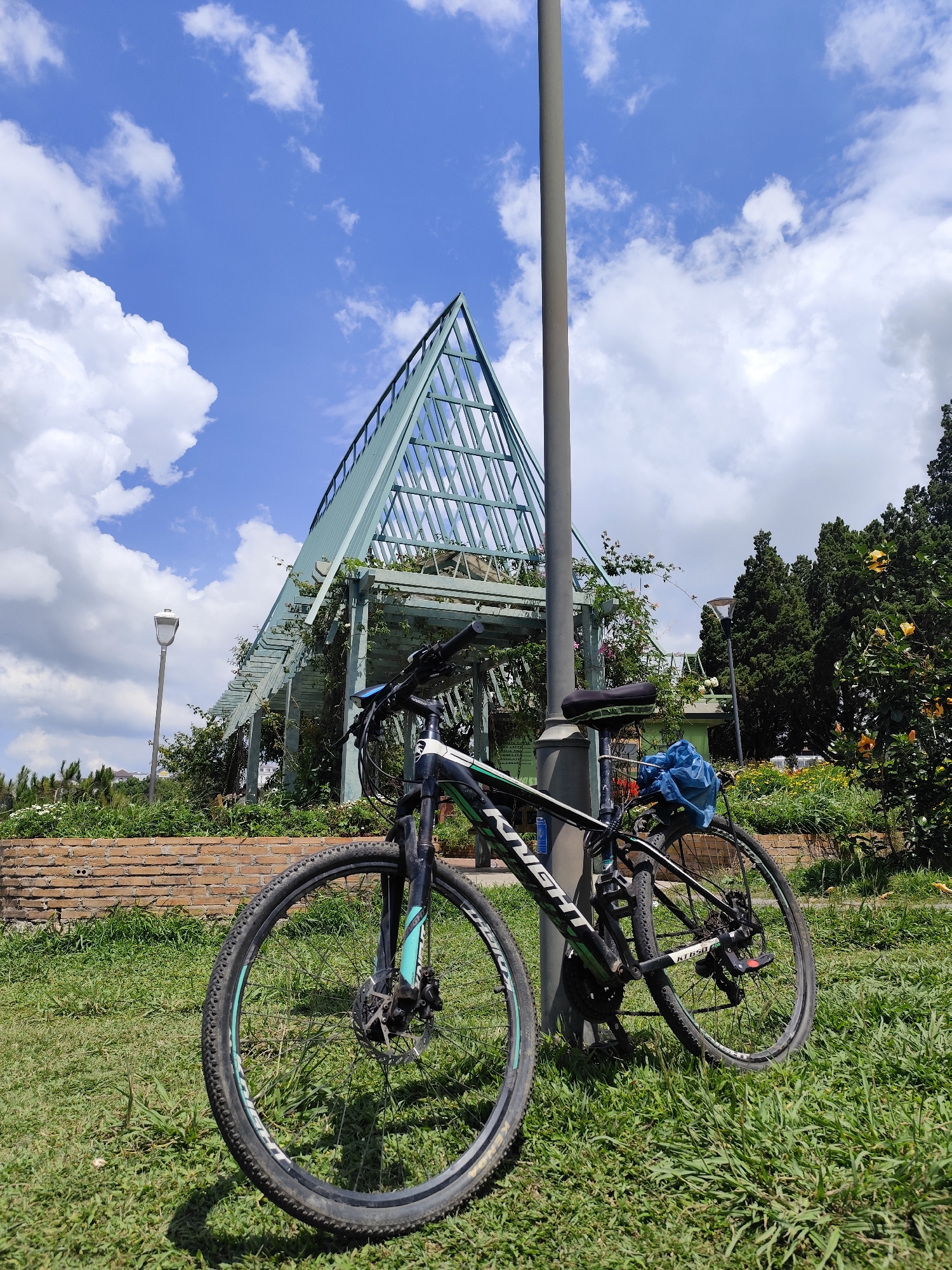 A bicycle leans against a lamppost in a park in Da Lat, Vietnam. The bicycle is black with green and white accents. A blue plastic bag hangs from the bicycle seat. Behind the bicycle is a metal pergola structure with a peaked roof, covered in vegetation. A brick wall is partially visible behind the pergola. To the right, there are trees and flowering bushes. The ground is covered in grass. The sky is blue with scattered clouds.