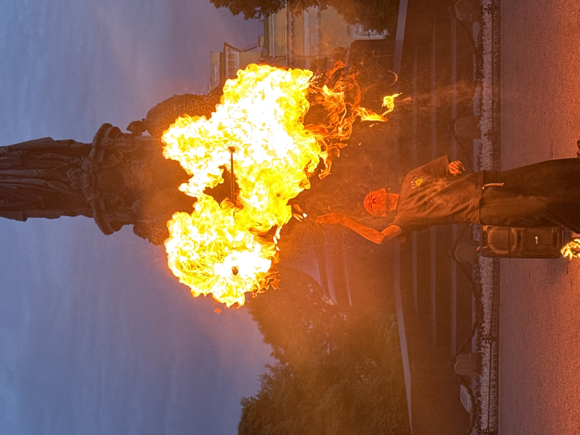 In Saint Petersburg, Russia, a performer is seen in front of the monumental Catherine the Great statue at Nevsky Prospekt. The performer, a young man wearing a dark t-shirt and dark pants, is in the process of breathing fire, creating a large, dynamic plume of flames that dominates the center of the image. His face is illuminated by the intense light of the fire, and his eyes are closed as he exhales. One arm is raised, holding a fiery object. Behind him, the imposing bronze statue of Catherine the Great stands atop a grand pedestal. The background also includes trees, a building, and the dusky twilight sky. The overall scene captures a moment of dramatic performance in a historical urban setting.