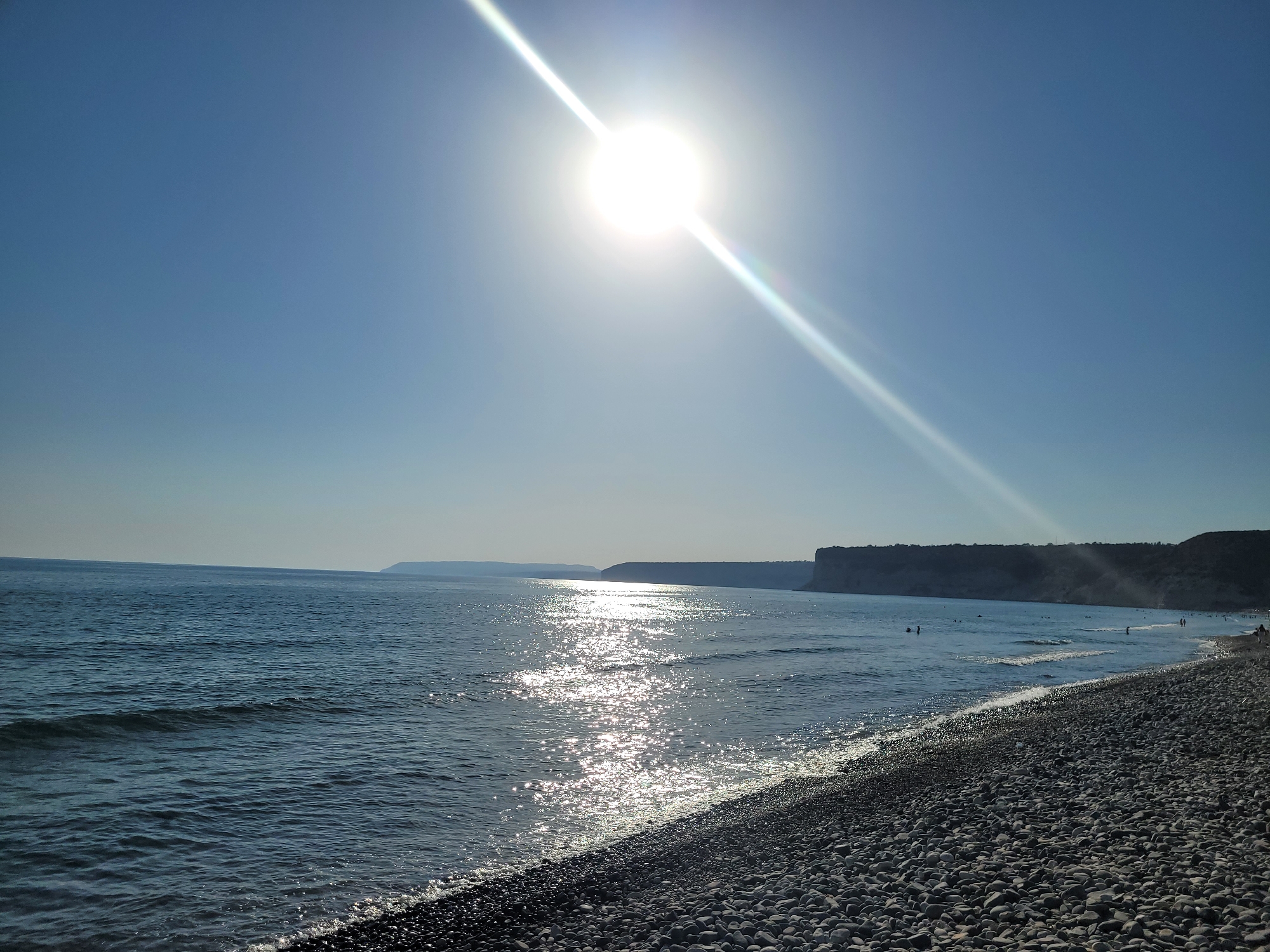 The image depicts a sunny day at a pebble beach in Episkopi, Cyprus. The sun is high in the clear blue sky, creating a bright glare that reflects off the gentle waves of the Mediterranean Sea. The shoreline is covered in smooth, grey pebbles. In the distance, cliffs rise along the coast, and a few people can be seen swimming and walking along the beach, enjoying the sea and sun. The overall atmosphere is serene and tranquil.