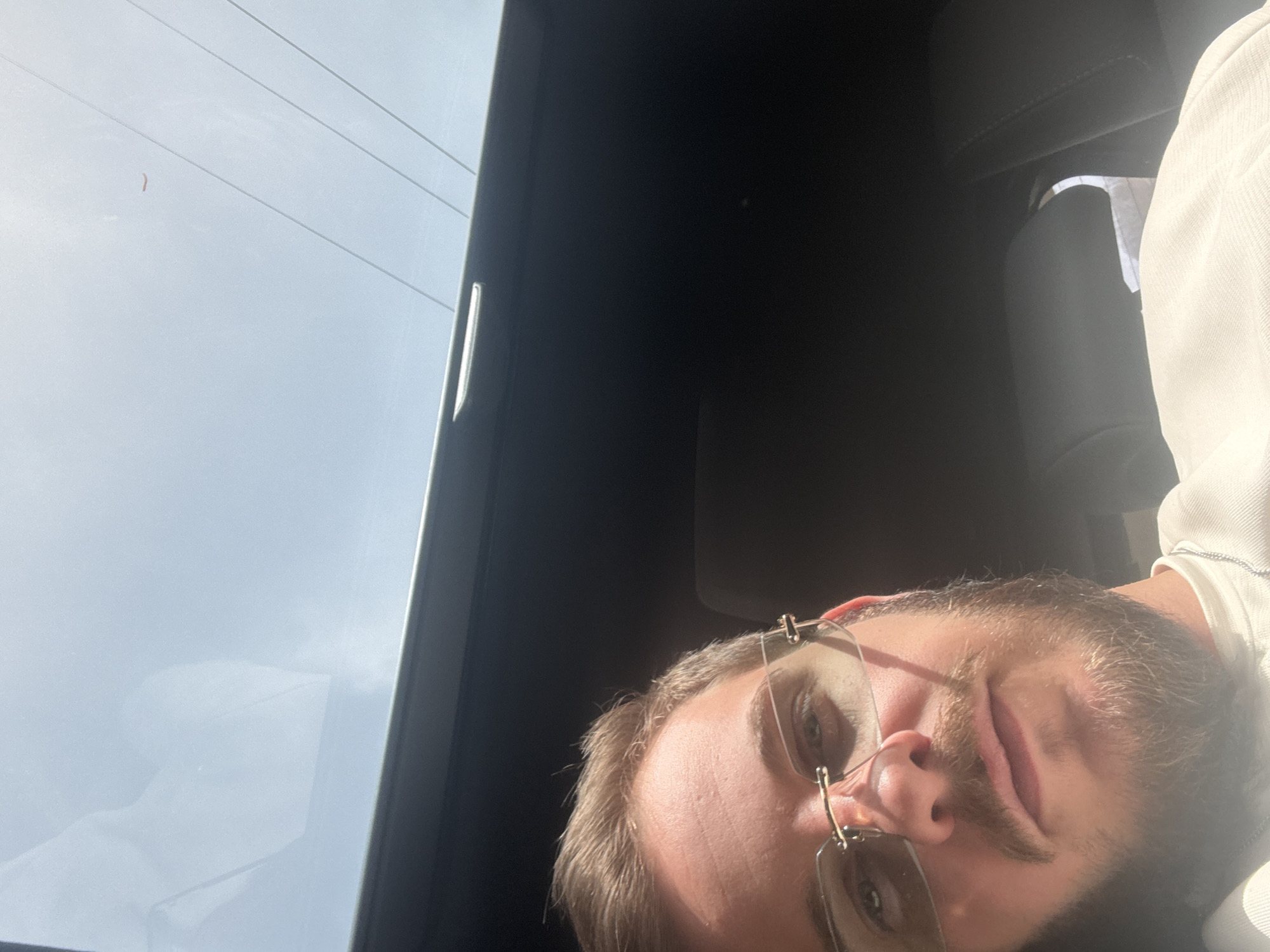 The image shows a selfie taken from inside a vehicle. The person in the image is a Caucasian male with blond hair and a beard, wearing glasses. He is positioned in the lower right corner of the frame. The interior of the car is visible, with a dark-colored dashboard and a portion of the seat. Outside the window, the sky is visible with power lines. The exact location in the city of Полатен, Bulgaria cannot be determined from the provided image.