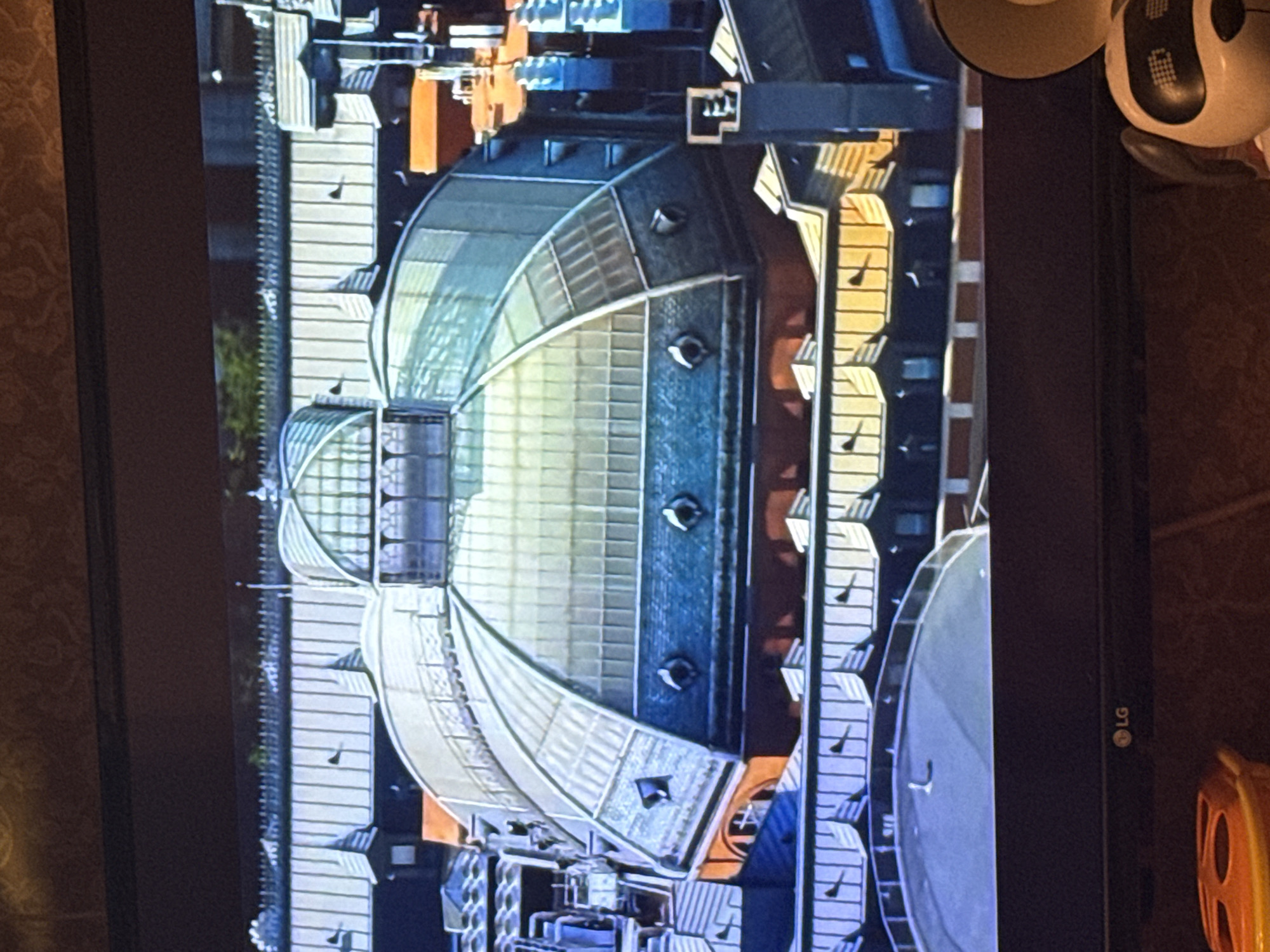 The image is a screen capture of a television displaying an aerial view of a building complex in Moscow, Russia. The architecture features a series of interconnected structures with glass-covered roofs in a semi-circular or fan-like arrangement. These glass roofs are segmented into rectangular panels. Adjacent to these structures are taller buildings with repetitive architectural details, possibly residential or office buildings. The colors are predominantly cool tones with a mix of blues, grays, and hints of orange. The foreground includes a portion of the television's bezel, and the LG logo is visible in the lower right corner of the TV. There is an unknown off-white object with black details visible at the top right of the screen.