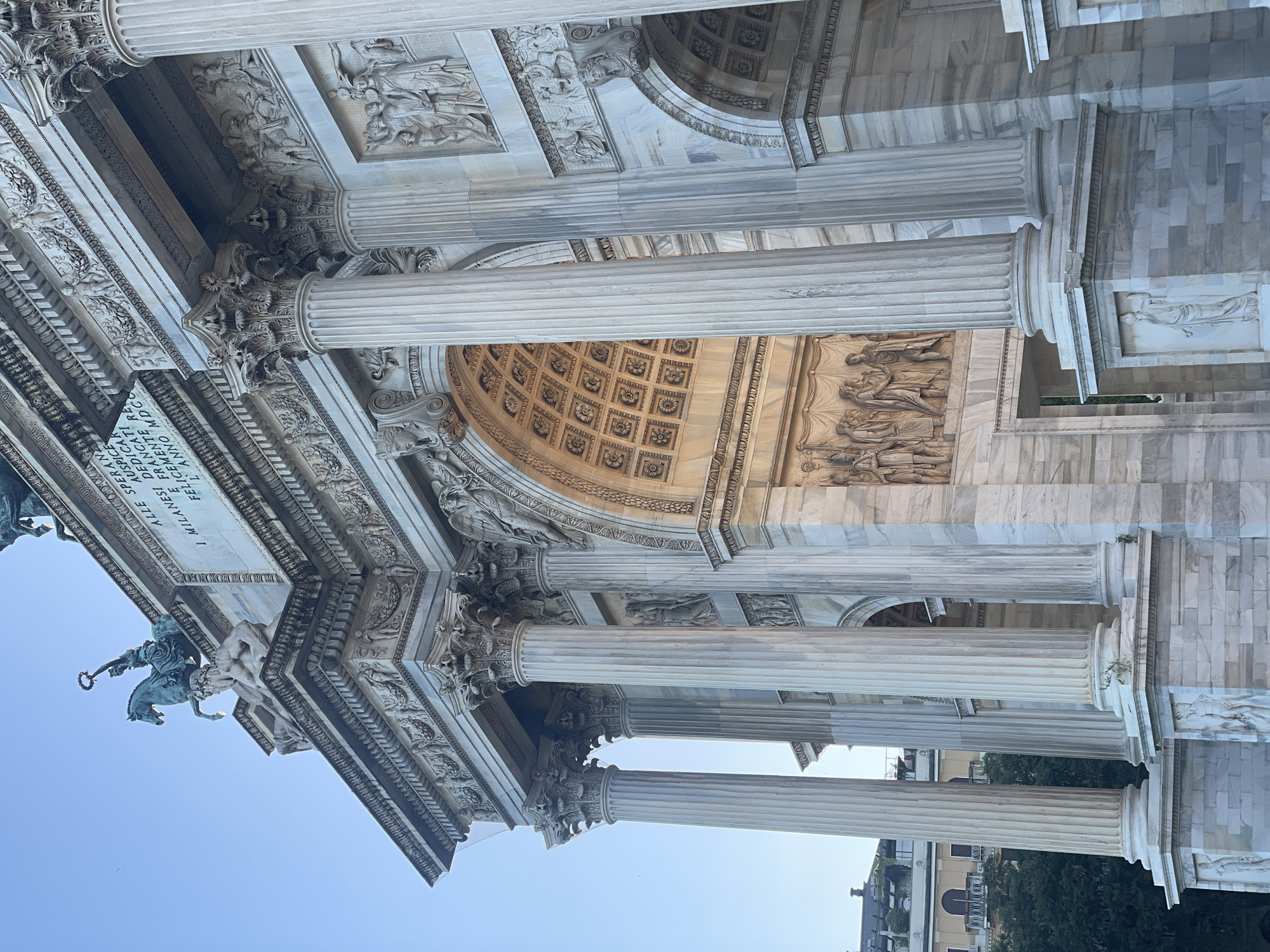 The image shows a detailed view of the Arco della Pace (Arch of Peace) in Milan, Italy. The arch is constructed of white marble and features intricate sculptures and relief carvings. Several fluted Corinthian columns are visible, supporting the entablature and pediment. On top of the arch, a quadriga (a chariot drawn by four horses) is depicted, though it is partially out of frame. The central archway is visible, revealing a coffered ceiling inside. Relief sculptures adorn the spandrels of the arch and the frieze above. The sky is clear and a light blue. In the background, some trees and buildings can be seen, hinting at the urban setting of the monument. The overall impression is one of grandeur and historical significance.