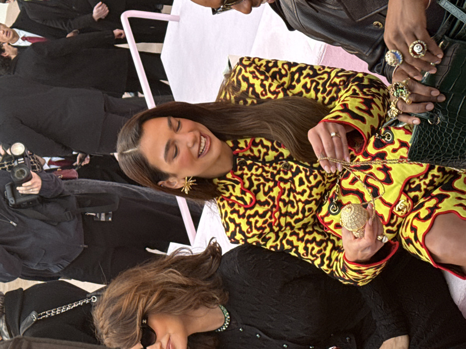 The image captures a close-up, medium shot of a high-profile indoor event, specifically the Chanel Spring/Summer 2024 Haute Couture show at the Grand Palais Éphémère in Paris, France.
Three individuals are seated on a light pink tiered bench or seating structure in the foreground. In the center, singer Dua Lipa is prominently featured, smiling with her head tilted down and to her left. She wears a vibrant yellow suit jacket adorned with a black and red abstract, leopard-like pattern and red scalloped trim. Her long, dark brown hair is parted down the middle, and she wears prominent gold starburst-design earrings. Both hands hold a small, spherical, intricately detailed gold handbag with a gold chain strap. A ring is visible on her left ring finger.
To Dua Lipa's left, actress Penélope Cruz is partially visible in profile, wearing dark sunglasses with a Chanel logo on the temple. She has wavy brown hair and is dressed in a black textured cardigan with a dark green beaded necklace visible underneath. A black handbag with a silver chain strap hangs over her shoulder.
On Dua Lipa's immediate right, the hands and partial shoulder of a third individual are visible. This person wears a black leather jacket and numerous large, ornate rings on their fingers, including one with a green stone and another with a blue and white design. Their hands rest on a dark green, crocodile-embossed handbag.
The background is blurred, showing several standing figures, including a photographer holding a professional camera with an attached flash unit. Other individuals appear to be in dark suits, some with red ties. A light pink railing or barrier is visible in the mid-ground.