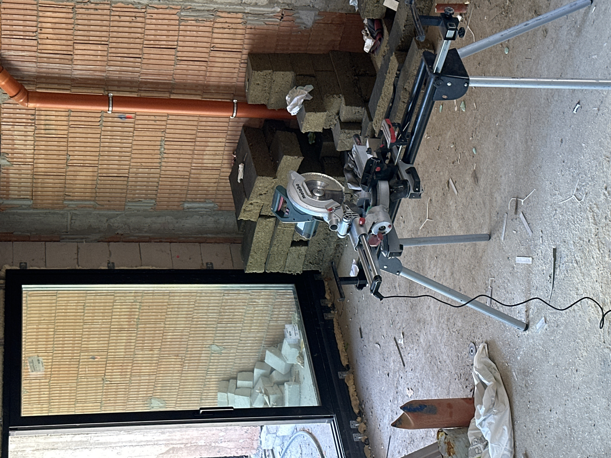 This image captures a construction site in Stara Zagora, Bulgaria. The scene is an unfinished interior room with exposed brick walls and a large glass sliding door revealing a pile of concrete blocks outside. In the foreground, a compound miter saw is set up on its stand, with its power cord running across the dusty concrete floor. Several stacks of dense, dark grey insulation material are visible in the middle ground, along with a few white cloths and some construction debris. An orange pipe runs vertically along the brick wall in the background. The overall impression is one of active construction work in progress.