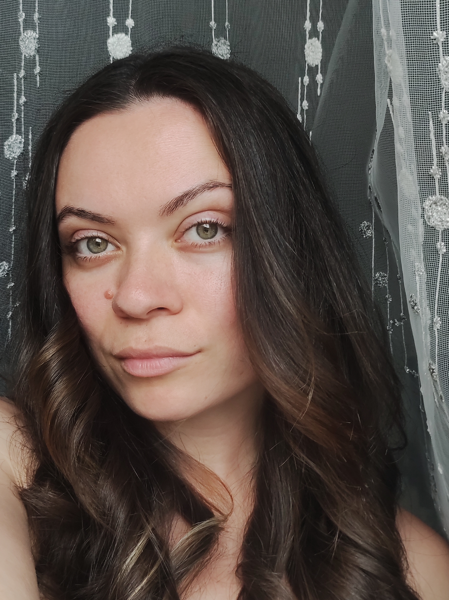 A close-up shot depicts a woman with long, wavy brown hair. Her eyes are light green. She has a skin tag near her nose. The background appears to be a textured curtain.