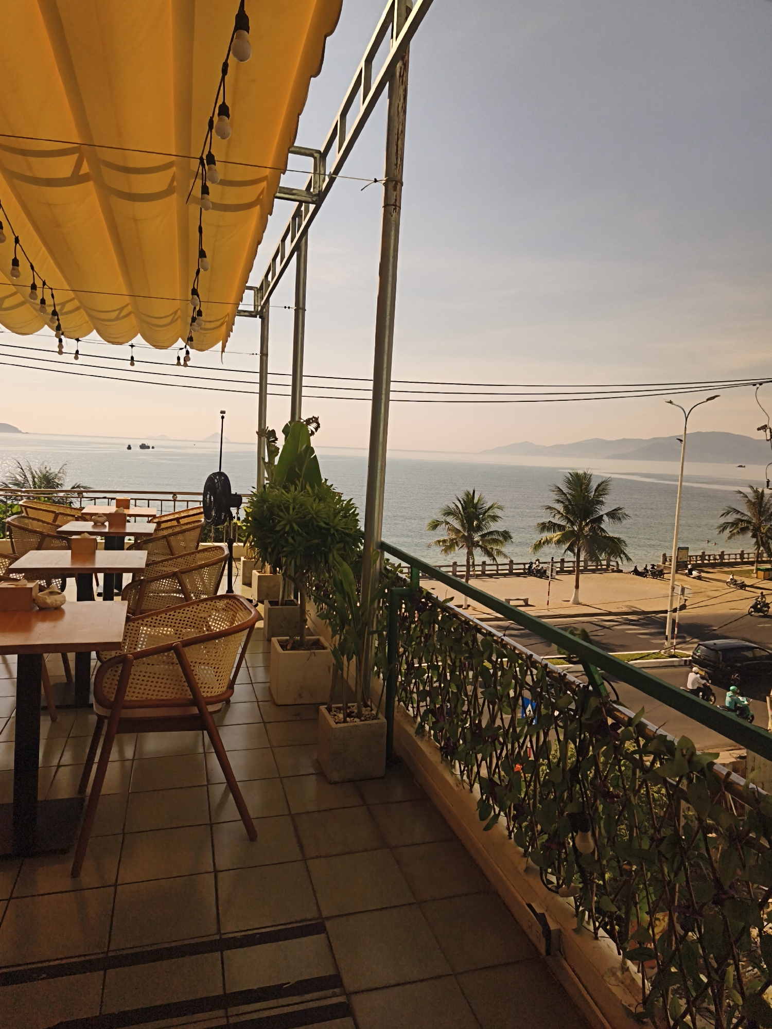 The image shows a view from the balcony of a restaurant in Bac Nha Trang Ward, Vietnam. The balcony is furnished with several tables and chairs, some of which are made of rattan. A yellow awning covers part of the balcony, and strings of lights are draped across it. In the foreground, there are potted plants and a black fan. The balcony overlooks the sea, with a beach, palm trees, and mountains visible in the distance. There are also some boats on the water and a street with a few vehicles. The time of day appears to be late afternoon or early evening, as the sun is low in the sky and casting long shadows.