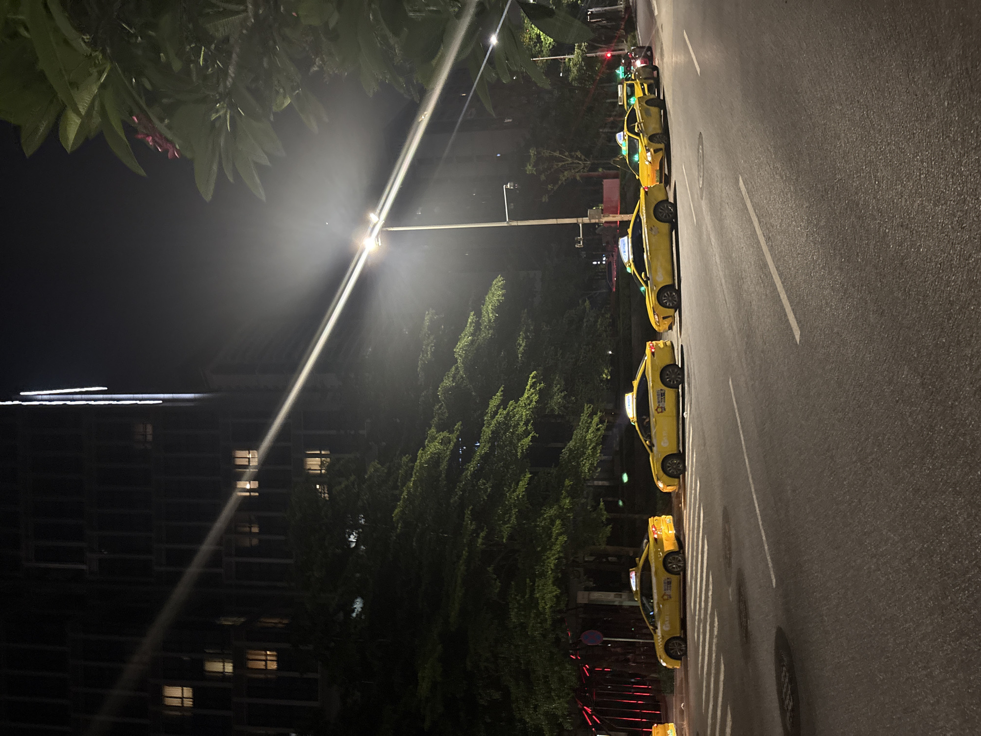 A row of yellow taxis are parked on the side of a street at night. The taxis are facing towards the left of the frame and appear to be waiting for passengers. The street is paved with asphalt and has white lane markings. Trees line the street, and a building is visible in the background on the left. The building has many windows, some of which are lit. A street lamp illuminates the scene, casting a bright light and creating a lens flare. The sky is dark, indicating it is nighttime. The image is taken from a slightly elevated perspective, looking down at the taxis and the street. There are no people visible in the image. The overall scene suggests a quiet urban environment at night, possibly in a commercial or residential area. There is some text visible on the taxis, but it is too small and blurry to read. The image is oriented vertically.