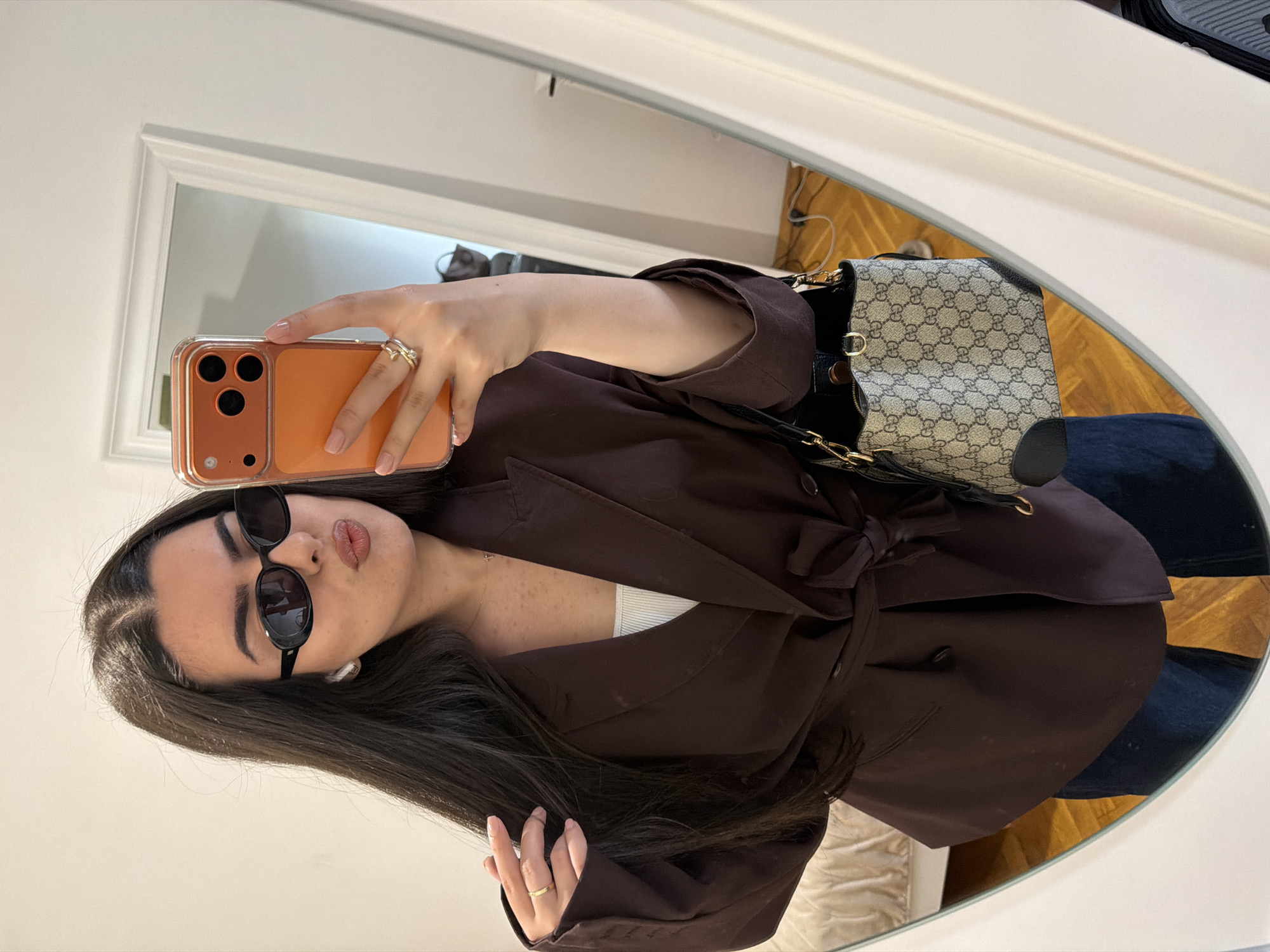 A woman is taking a selfie in a mirror, presumably indoors. She is wearing dark sunglasses, a brown belted jacket over what appears to be a white top, and is holding an orange iPhone. She has dark hair and is wearing rings on both hands. Behind her, reflected in the mirror, is a beige patterned handbag with a black strap, and a glimpse of what may be a room interior. The specific location in Sofia, Bulgaria, cannot be determined from the given visual information.