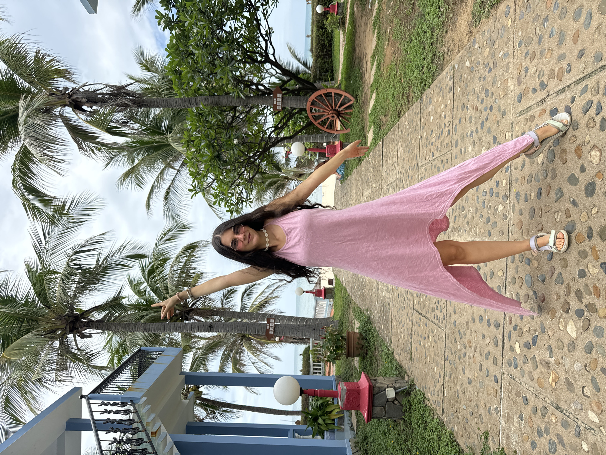A cheerful woman, the central subject of the image, poses dynamically outdoors, extending her arms and raising one leg, creating an impression of joy and freedom. She wears a flowing pink sleeveless dress, sunglasses, a shell necklace, and bracelets on her right wrist. Her long, dark hair cascades down her back.
She stands on a path paved with embedded pebbles, characteristic of a garden or resort walkway. The setting is lush and tropical, dominated by numerous tall palm trees and various green plants and bushes lining the path. In the mid-ground, a red wooden wagon wheel is affixed to a tree, adding a rustic decorative element, and red-based spherical white lamps are visible along the path.
In the background, the sparkling blue ocean meets the horizon under a partly cloudy sky, confirming a coastal location. To the left, a multi-story building with blue railings and white accents suggests resort architecture. The overall scene conveys a bright, pleasant daytime atmosphere, likely in a tropical vacation spot. While there is a white sign with text on one of the palm trees, its content is unreadable. Given the context, the location is Mũi Né, Vietnam.