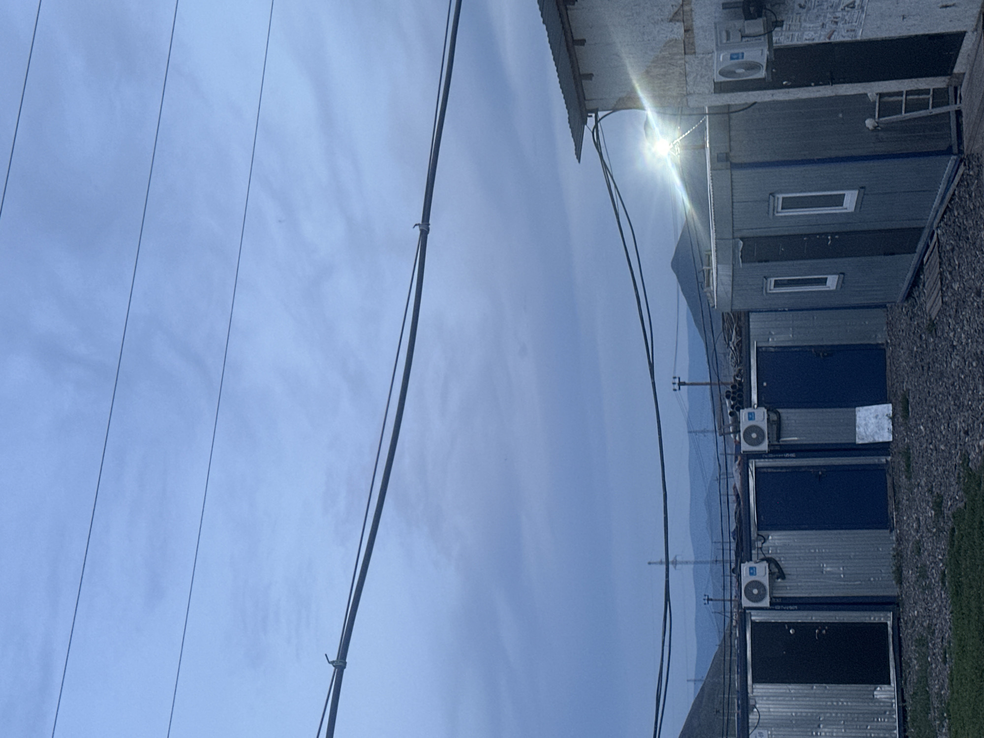 The image depicts a row of modular buildings, likely shipping containers repurposed as structures, in Найырал, Russia. The scene is set outdoors during what appears to be late afternoon or early evening, with a bright sun glinting off the corrugated metal siding and roof of one building. The sky is predominantly a hazy, pale blue with some wispy clouds, suggesting a partly cloudy or dusty atmosphere.
The modular buildings are stacked or placed adjacent to each other, featuring dark blue doors and rectangular windows. Several of the structures are equipped with outdoor air conditioning units, indicating a need for climate control. Power lines crisscross the sky in the foreground, adding to the utilitarian aesthetic of the setting. In the background, a range of distant, hazy mountains is visible, suggesting a somewhat rural or mountainous location. The ground around the buildings is a mix of gravel and patchy green grass. No people are visible in the frame, and there are no discernible emotions conveyed. A few labels or signs with text are visible on the buildings, but they are too small and indistinct to be read.