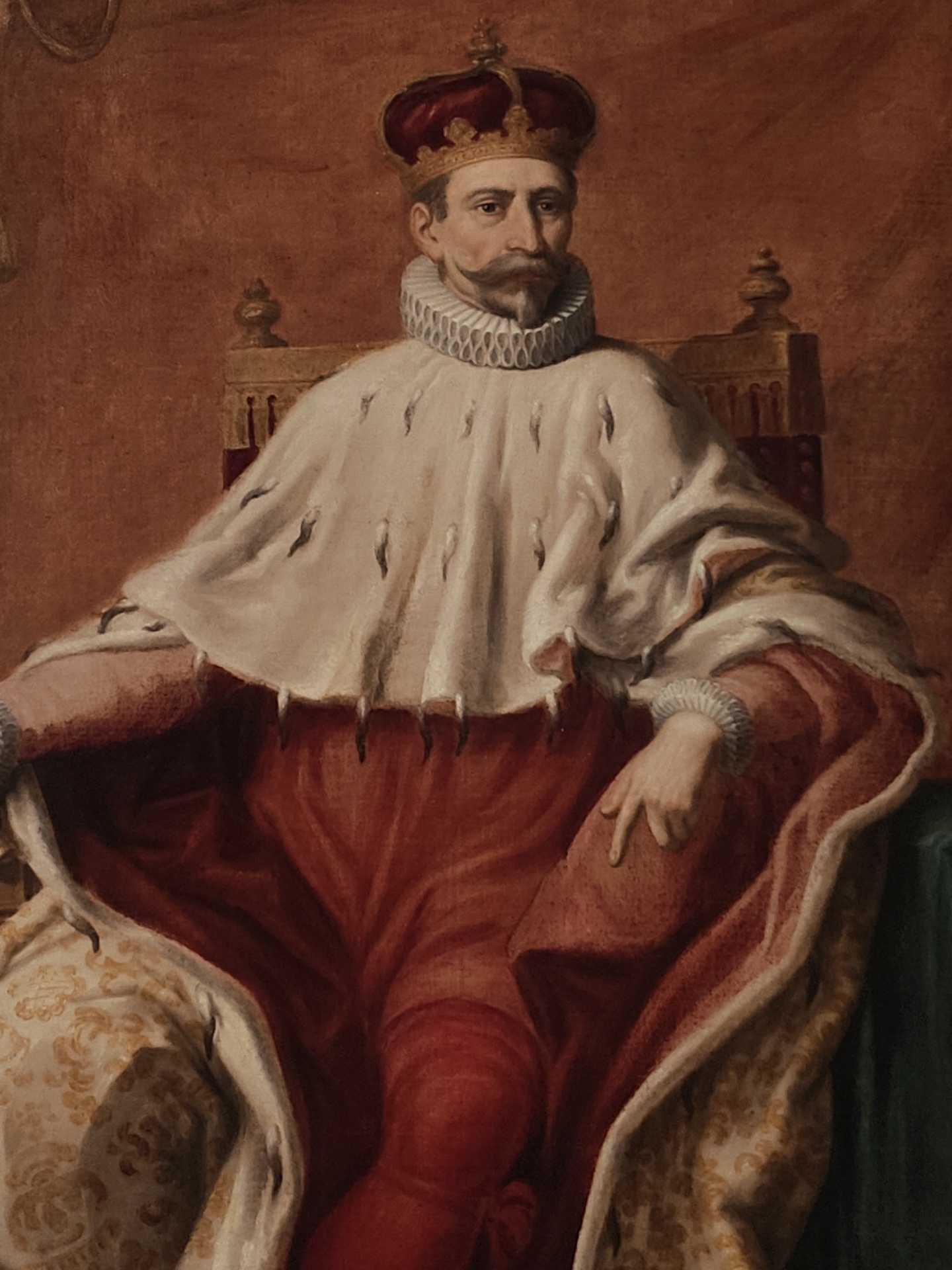 The image is a portrait of a man, possibly a king or nobleman, seated on a chair. He is wearing a crown, a ruffled collar, and a regal robe with ermine trim. The robe is predominantly red, with sections of white and gold brocade. He has a mustache and a goatee. His right hand rests on his leg, while his left is partially visible in his lap. The background is a plain, reddish-brown color. The chair has gold finials. There is no direct information in the image specifying the location in Genoa, Italy.