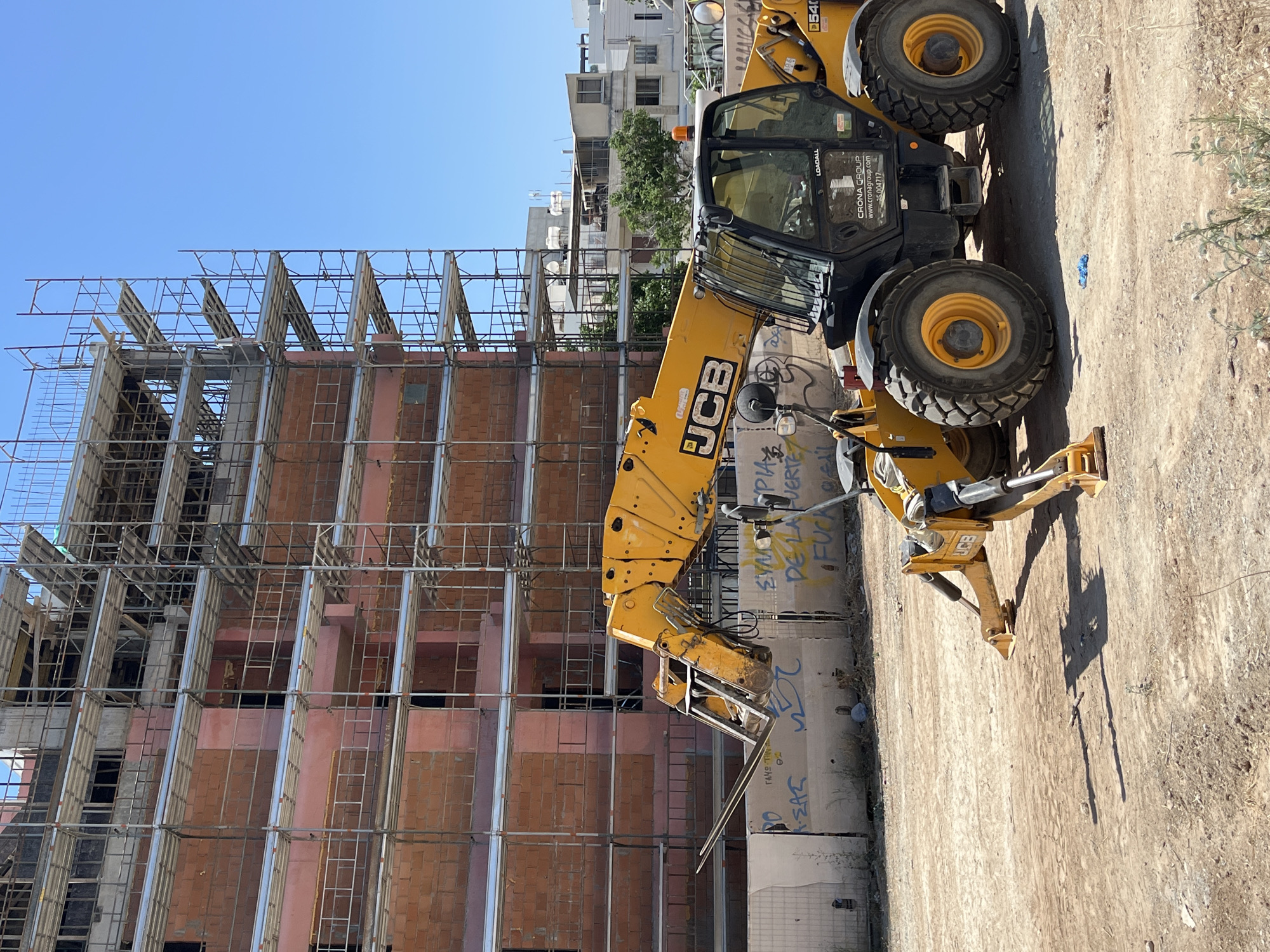 In Limassol, Cyprus, a construction site is depicted with a partially built building covered in scaffolding. A yellow telescopic handler, identified by the "JCB" logo, is parked on a dirt lot in front of the building. The handler has its forks extended and positioned low to the ground. Graffiti adorns a concrete barrier separating the lot from the building's foundation. The sky is clear and blue.