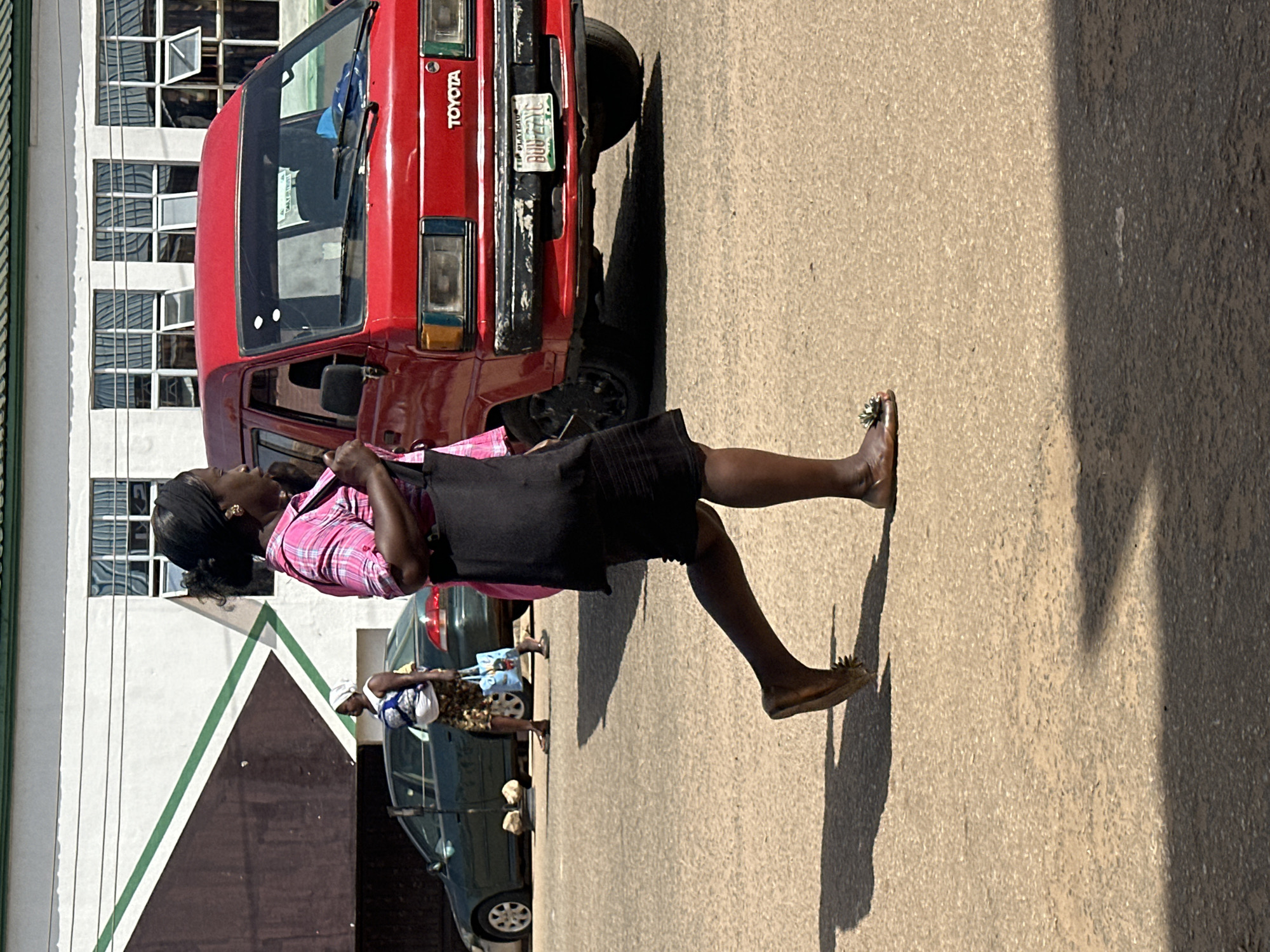 A woman in a pink checkered shirt and black skirt walks on a paved street in Bukuru, Nigeria. She carries a black bag over her left shoulder and wears sandals. Behind her, a red Toyota van is parked. To the left of the van, a dark green car is visible, and another woman wearing a white headscarf and blue patterned skirt stands with a light blue tote bag. The background shows a building with white walls and multiple windows with white frames. The scene is illuminated by sunlight, casting shadows on the street.