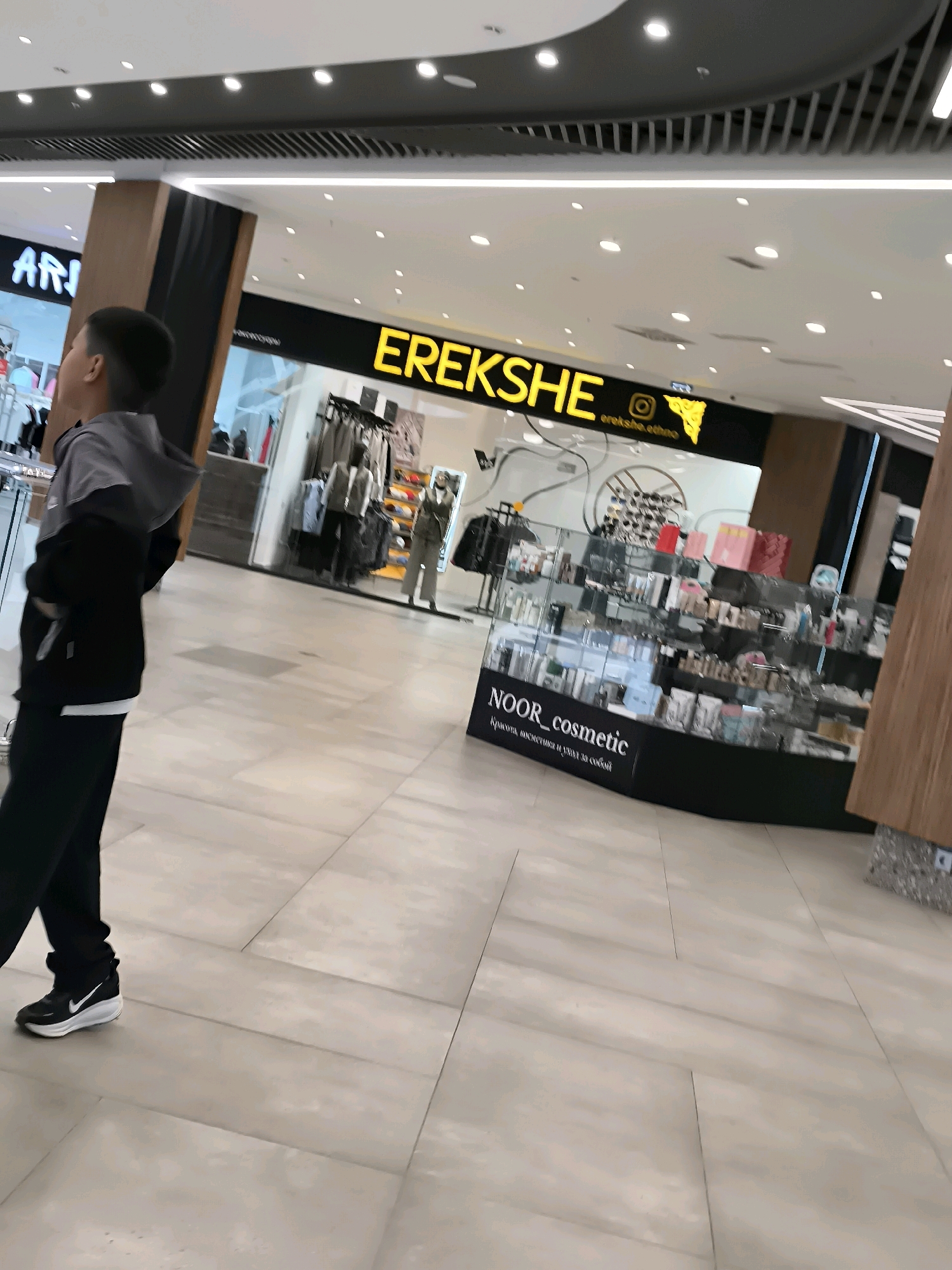 The image shows a young boy walking through a modern shopping mall. The boy is wearing a grey hooded jacket over a black shirt and black pants, with black Nike sneakers. In the background, there are shops, including one with a yellow sign that reads "EREKSHE" and another with a sign for "NOOR cosmetic". The mall features tiled floors and a well-lit ceiling with circular lights and ventilation systems. The area visible appears to be part of a commercial district within a city, potentially Qarağandı city administration, Kazakhstan, based on the provided context, though the image itself does not explicitly state the location.