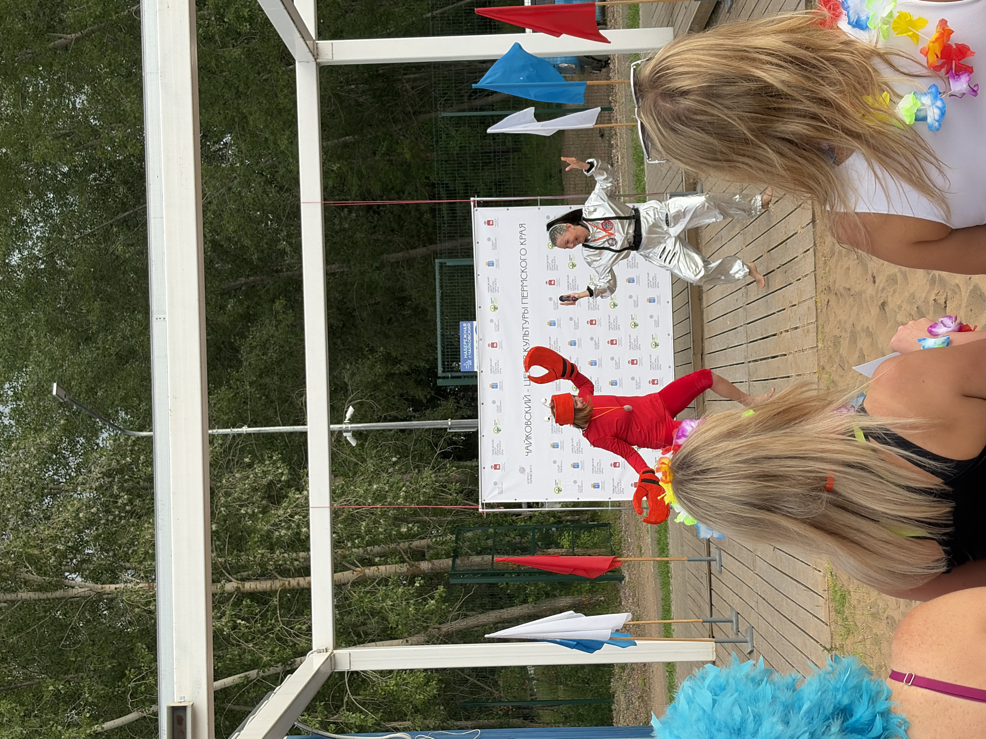 The image captures an outdoor event at the embankment of the city of Chaikovsky, Russia. In the foreground, two individuals are viewed from behind, one with a bright blue feathery headpiece and the other with blonde hair adorned with a floral lei. The main subjects are on a wooden stage in the mid-ground, facing away from the camera. One performer is dressed in a red costume with large red crab claws on their hands and a matching headpiece. The other performer is wearing a shiny silver, futuristic-looking jumpsuit. They are both actively engaged in a performance, with their arms outstretched and dynamic stances suggesting movement. Behind them, a large white banner is displayed, featuring text in Russian and various logos. Several flags, including white, red, and blue, are planted in the sand to the left of the stage. The background consists of trees and a fence, indicating an outdoor, possibly park-like setting. The overall atmosphere appears festive, suggesting a public event or entertainment on a beach or waterfront.