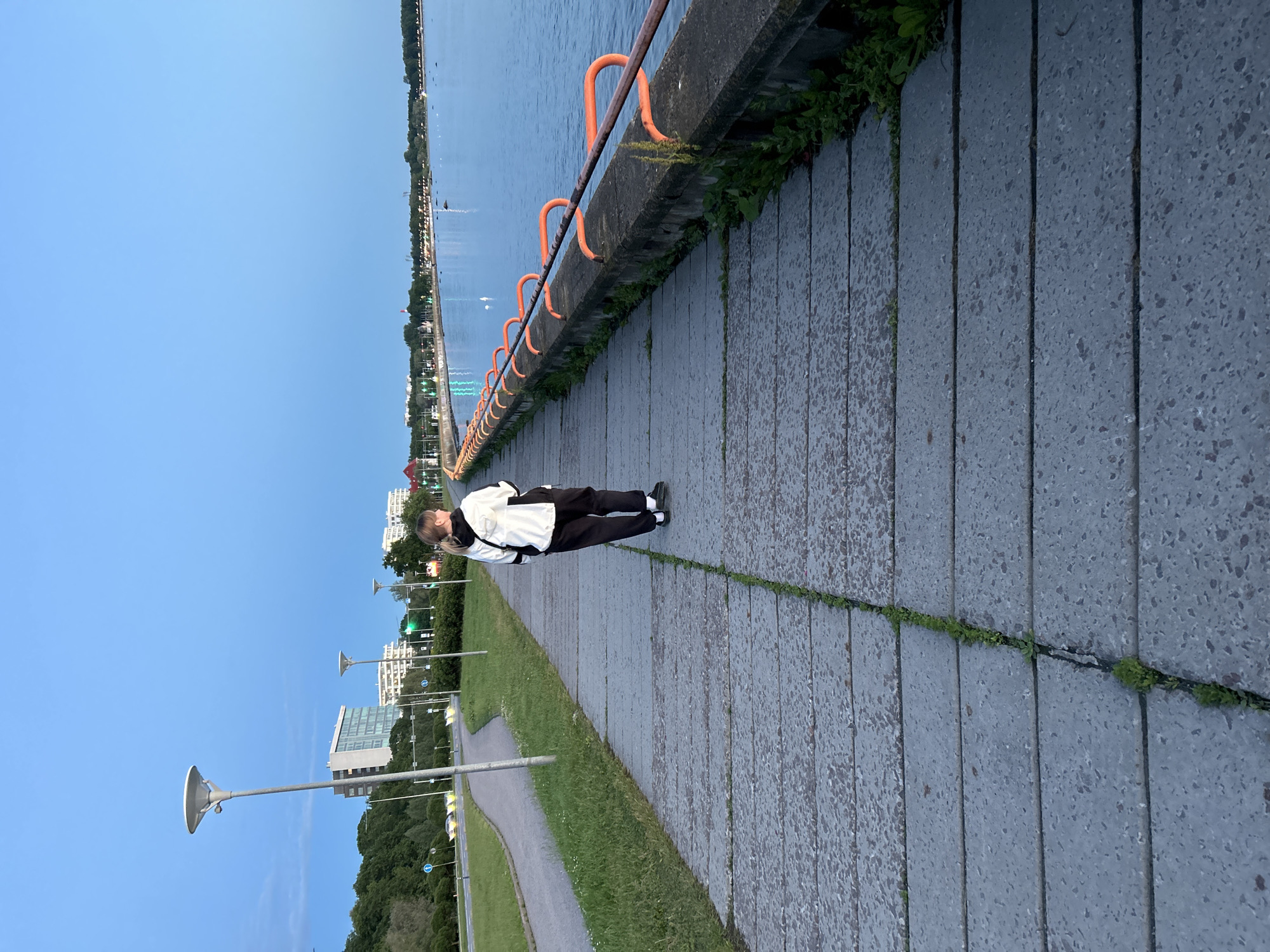A lone person walks along a concrete promenade next to a body of water. The person is wearing a black and white jacket and black pants, and is facing away from the camera, looking towards the water. The promenade is lined with an orange railing and is bordered by grass and trees on one side, and the water on the other. In the background, there are buildings and streetlights. The sky is a clear, pale blue. This scene is located in Tallinn, Estonia, on the Pirita promenade, a popular waterfront area known for its views of the Baltic Sea and its recreational facilities.