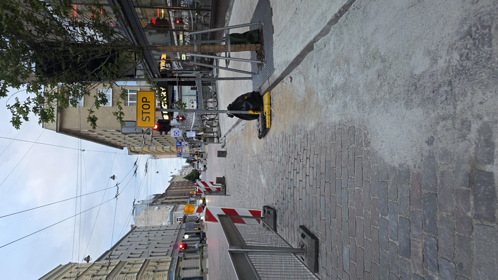 The image depicts a street scene in Riga, Latvia, likely during the daytime given the lighting conditions, though the sky appears overcast. The street is paved with cobblestones in the foreground, transitioning to a lighter colored surface, possibly concrete or sand, further down the lane. Temporary metal barriers with red and white striped markers are present, indicating ongoing construction or roadwork.
A prominent yellow "STOP" sign is visible on a pole, accompanied by a red traffic light. A large black trash bag is placed on a yellow base on the sidewalk, near a young tree that is supported by metal scaffolding. In the background, several buildings line the street, exhibiting varied architectural styles typical of European cities. There are also overhead wires, suggesting the presence of trams or other electric transport.
A few vehicles are visible in the distance, including cars and what appear to be bicycles parked on the side. The overall impression is one of a working city with infrastructure maintenance in progress, creating a temporary disruption to the usual flow of traffic and pedestrian activity. There are no clearly discernible individuals in the foreground. The scene conveys a sense of urban functionality and development.