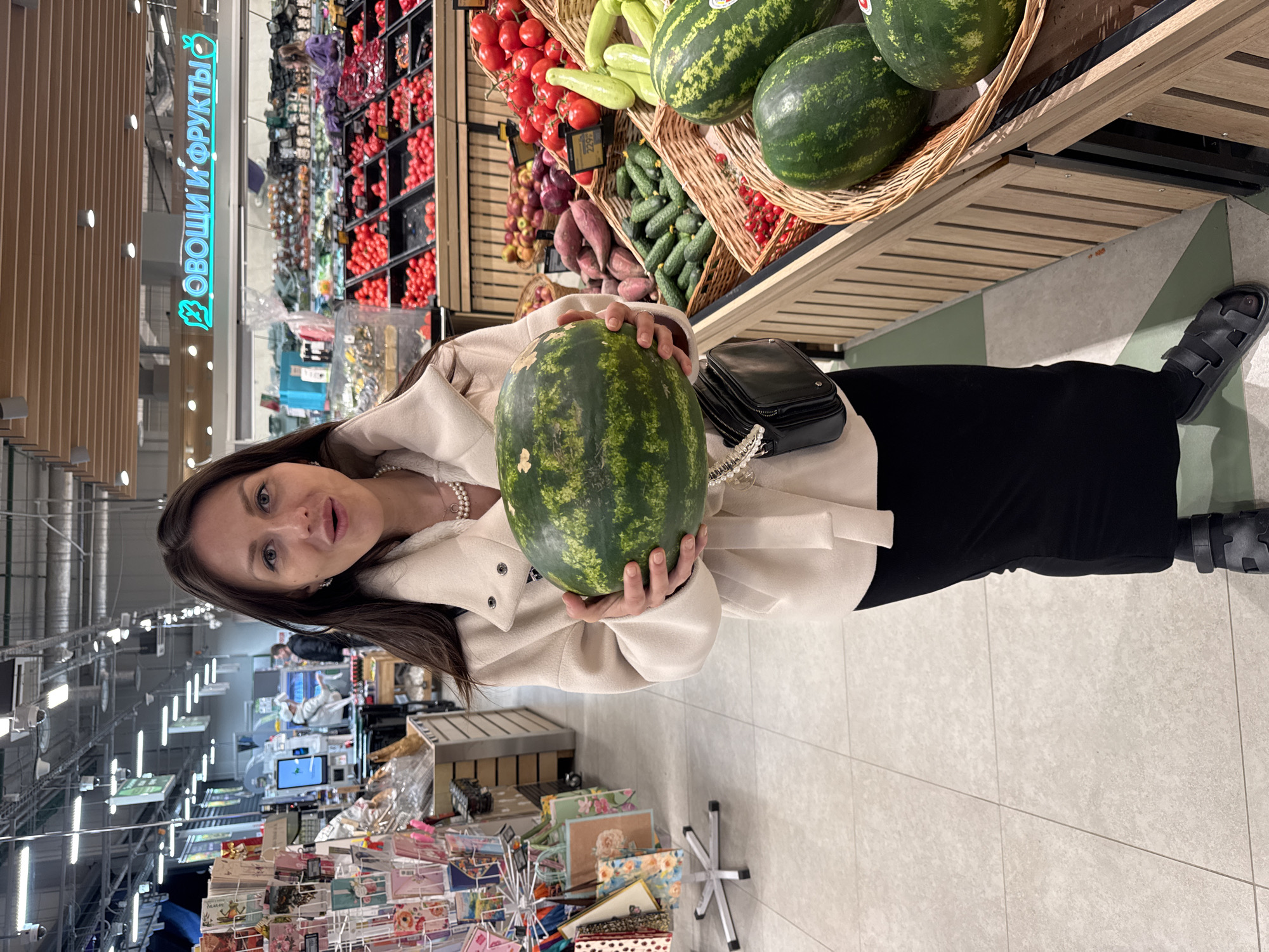 A woman holds a watermelon in a store, with a shelf of fruits and vegetables behind her, including tomatoes, cucumbers, and other watermelons in a basket. She is wearing a light-colored jacket, a black skirt, and sandals. Her hair is dark, and she is also wearing a pearl necklace. In the background, there are shelves of various products, and signs are visible in a foreign language. The floor is tiled, and the overall setting appears to be a supermarket or grocery store.