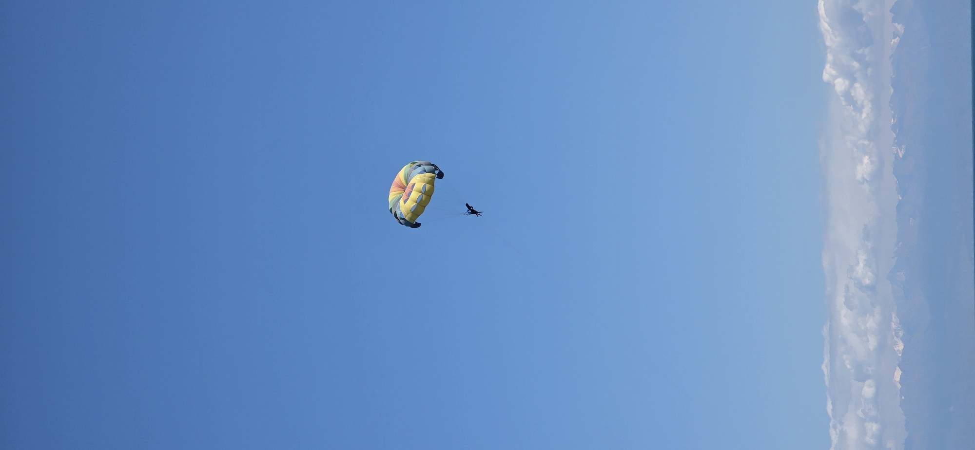 A brightly colored parasail, with stripes of yellow, red, green, and blue, is suspended against a clear blue sky. A person, appearing as a dark silhouette, is attached to the parasail by tow lines. The parasail is positioned in the upper middle section of the frame, slightly to the right. In the lower portion of the image, a distant, hazy mountain range can be seen, with a layer of clouds above them. A thin, dark horizontal line, likely representing the horizon or a body of water, is visible at the very bottom of the frame. The overall scene depicts an outdoor recreational activity in a scenic, possibly coastal or mountainous, environment. The location is specified as Bulan-Segettu, Kyrgyzstan.