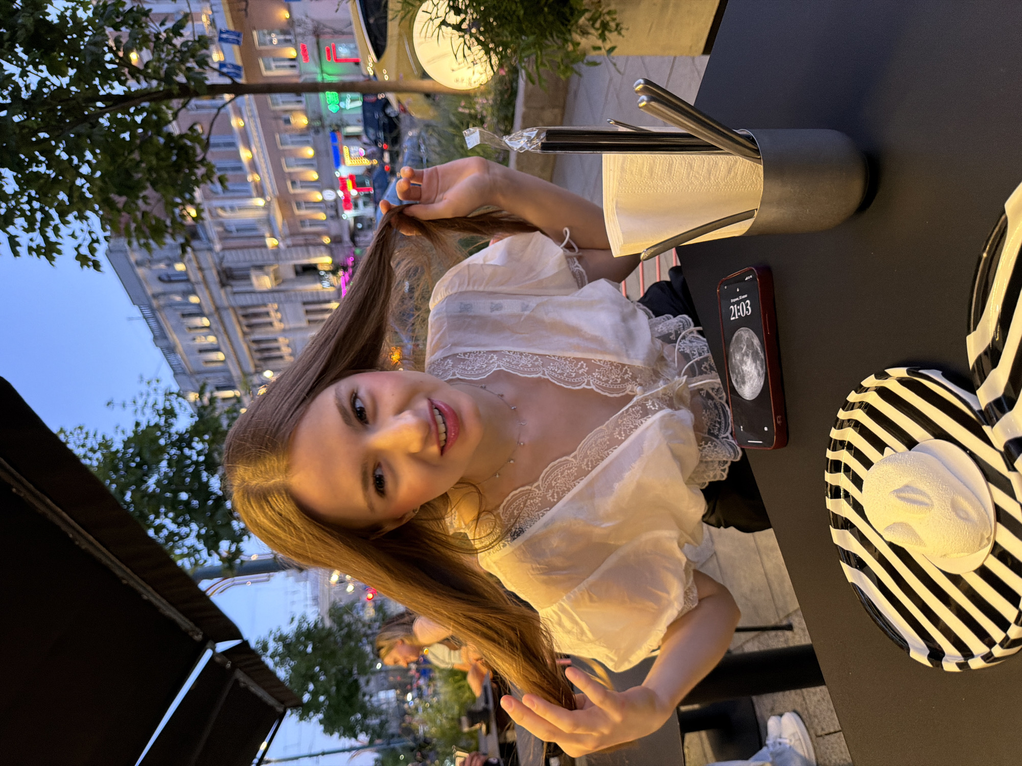 A young woman with long, brown hair is sitting at an outdoor table at dusk. She is wearing a white, lace-trimmed blouse and black pants. She is holding her hair in her hands and looking at the camera with a slight smile. On the table in front of her is a plate with a black and white striped pattern, a white, frosted dessert that resembles a mouse or other small animal, and a red smartphone displaying the time 21:03. To her right, there is a metallic cup holding napkins and utensils, and a glowing lamp with a decorative plant. In the background, there are buildings with illuminated windows and trees, suggesting an urban setting. The sky is a deep blue, indicating the time of day is evening. The image does not contain any explicit information or visual cues to confirm the location as Novaya Usman', Russia.