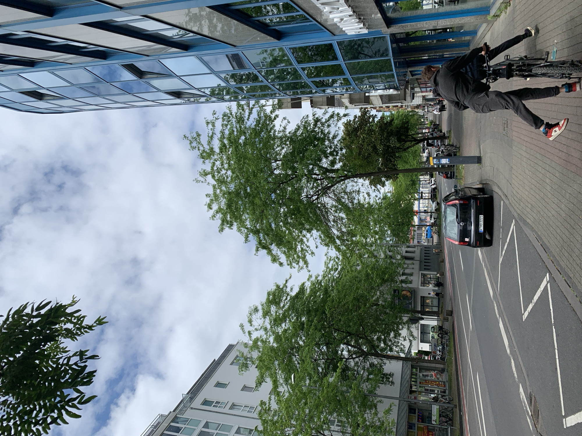 A daytime urban street scene is depicted, featuring a mix of modern and traditional buildings, green trees, and human activity. The sky is largely covered with clouds, suggesting an overcast or partly cloudy day.
In the immediate foreground, a person is riding a bicycle on what appears to be a sidewalk or designated bike path alongside the road. The cyclist is wearing a dark hoodie and dark pants, and is notable for their distinctive red and white athletic shoes. They are looking forward, actively engaged in cycling.
To the left, a prominent modern building features a blue structural frame and many reflective glass windows, which mirror the cloudy sky and surrounding trees. Further down the street, another white building with multiple rectangular windows is visible. The right side of the image shows a sidewalk and a street with a black car, identifiable by its license plate "K MO 2221", parked by the curb. The road surface has white lines, including a distinctive zigzag pattern.
Several green trees are interspersed along the street, their foliage providing a natural contrast to the urban architecture. In the background, other pedestrians can be seen walking on the sidewalk, and the facades of various businesses are visible. One partial sign in the background displays the text "WEIHETT". The scene captures a typical city environment during daylight hours.