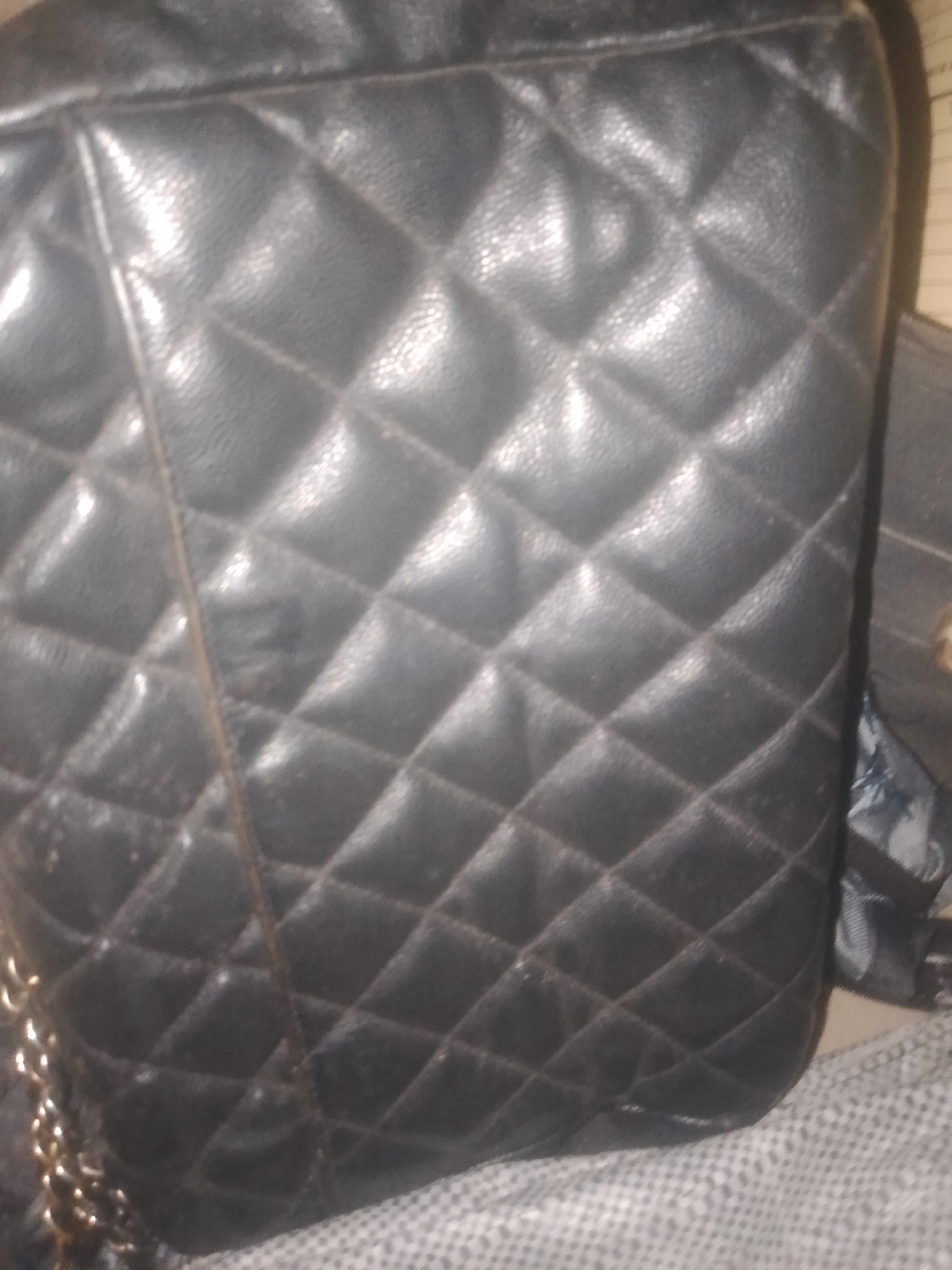 The image is a close-up, angled shot of a quilted black leather bag. The bag has a diamond-pattern stitching. A portion of a gold-colored chain strap is visible on the left side of the image. The bag is resting on a textured gray surface. The background is out of focus and appears to be a wall or fabric. There is no indication of the city of Jauro Yinu, Nigeria, or any specific location within it. The scene is an indoor still life of a handbag.