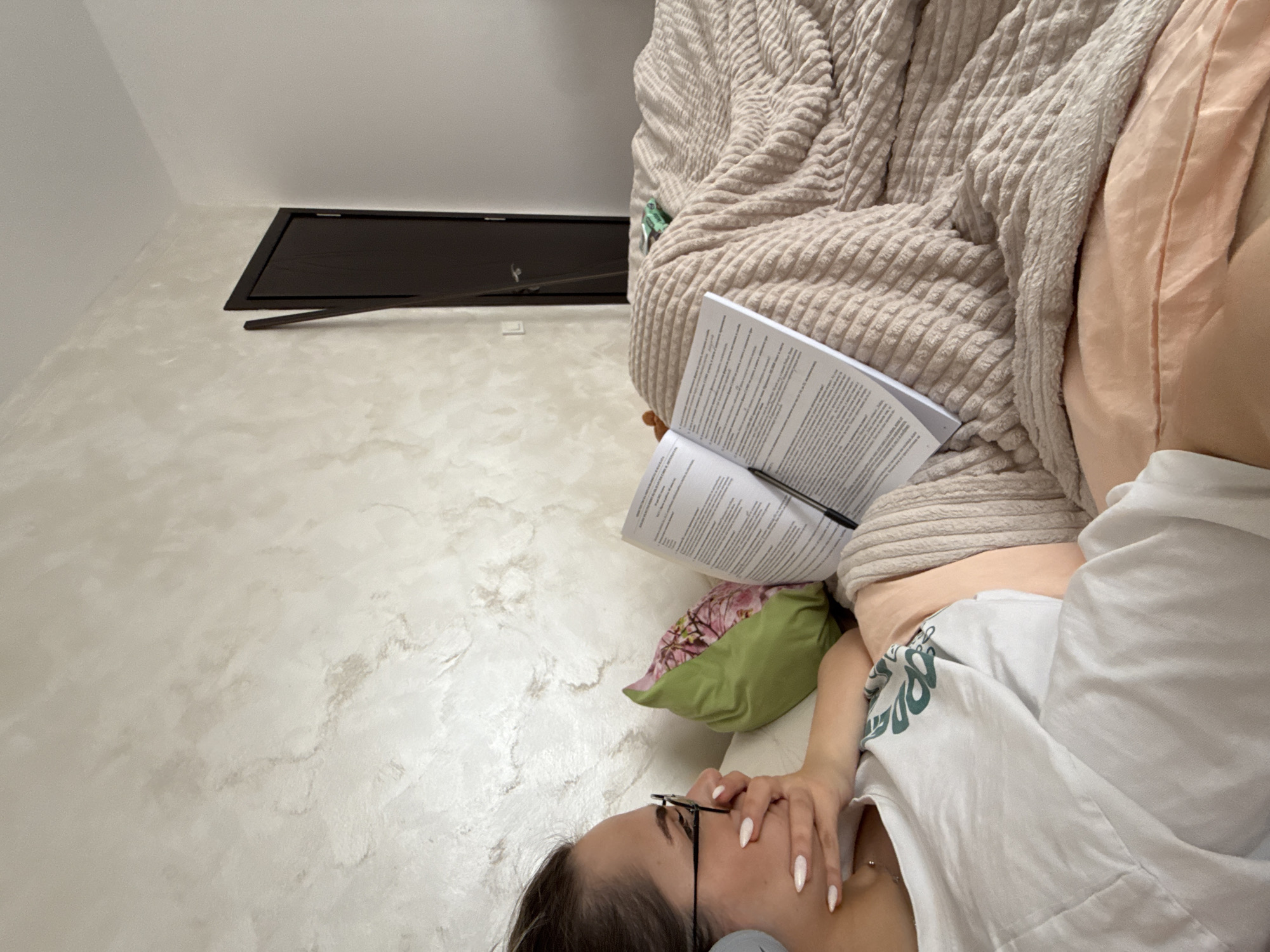 A woman lies on a beige corduroy beanbag chair in a room with a light beige, textured wall. She wears glasses, a white shirt with a green design, and has long white manicured nails. She rests her hand near her face. A green floral pillow supports a book, open to a page of text, with a black pen lying on top. A dark brown rectangular vent is visible on the wall in the background. The location is not evident from the provided image.