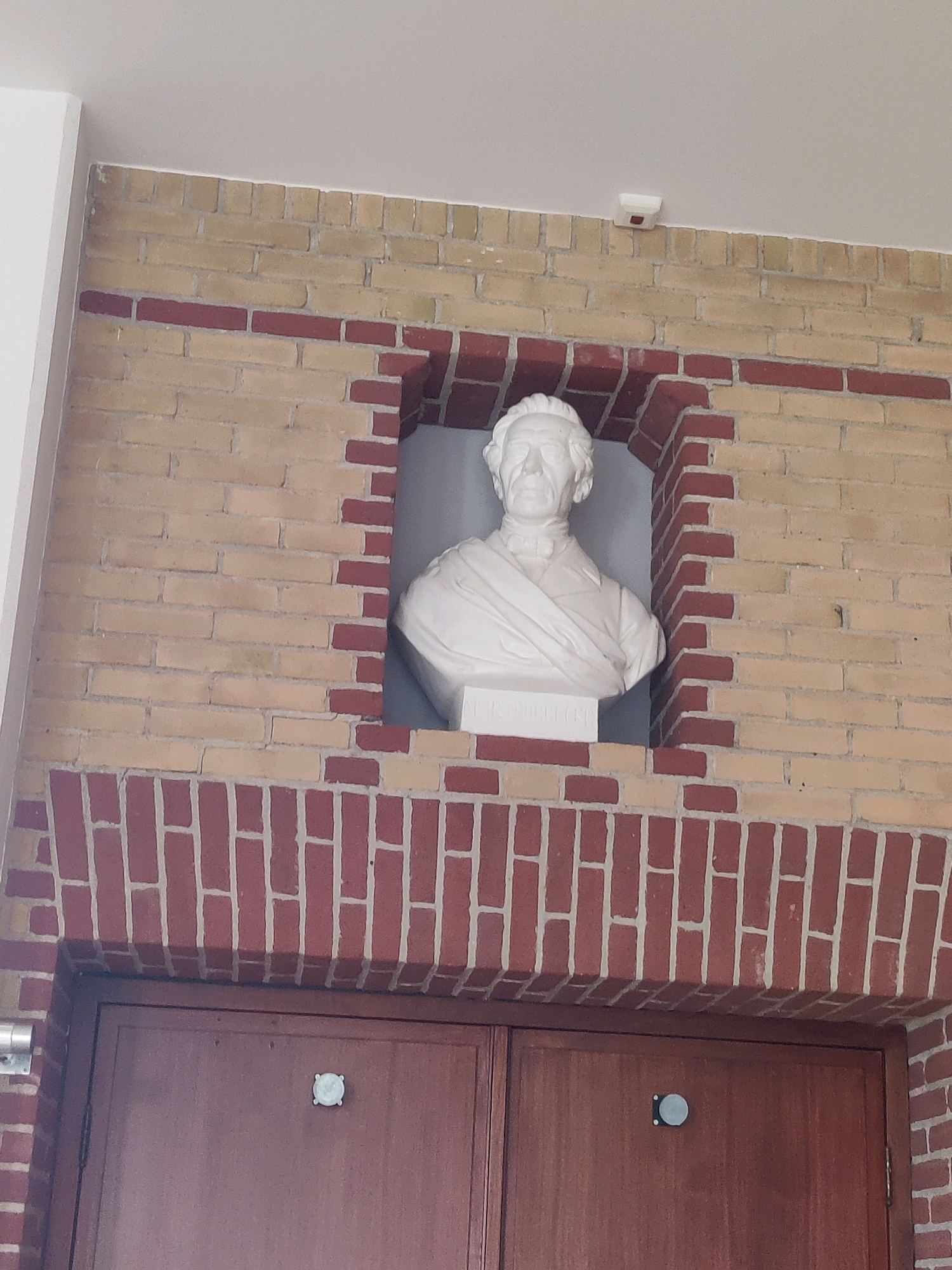 The image shows a white marble bust of a man, positioned in a recessed brick niche. The niche is framed by red bricks, with a lighter colored brick wall surrounding it. Below the niche are two large wooden doors. A small white and red device, possibly a smoke detector, is mounted on the wall above the niche. The bust depicts a man with short, wavy hair, a high forehead, and a stern expression. He is wearing a suit with a cravat, and a draped cloth over his shoulders. The bust is mounted on a white pedestal. The inscription on the pedestal is illegible in this image. This bust is located in the Willibrordusplein in Winterswijk, Netherlands, in front of the Willibrordus Church.