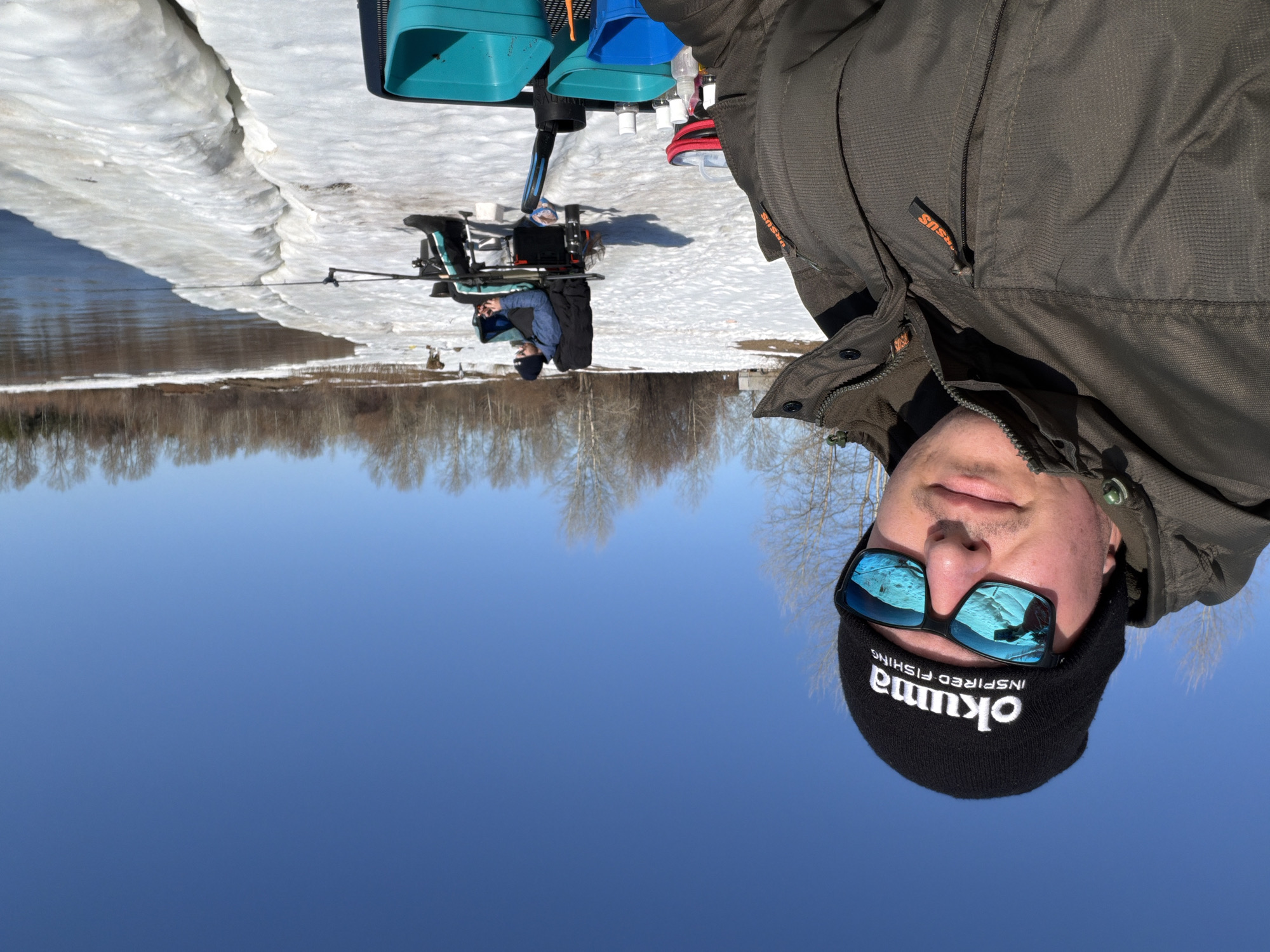 The image shows a man in a dark green jacket and black Okuma-branded beanie, wearing sunglasses, taking a selfie. The background reveals a winter fishing scene in Chaikovsky, Russia. A person is seen fishing on a snow-covered bank by a river or lake, with fishing equipment nearby. Trees line the opposite bank, and a clear blue sky is visible. There are patches of snow on the ground. The scene appears to be near a body of water surrounded by trees.