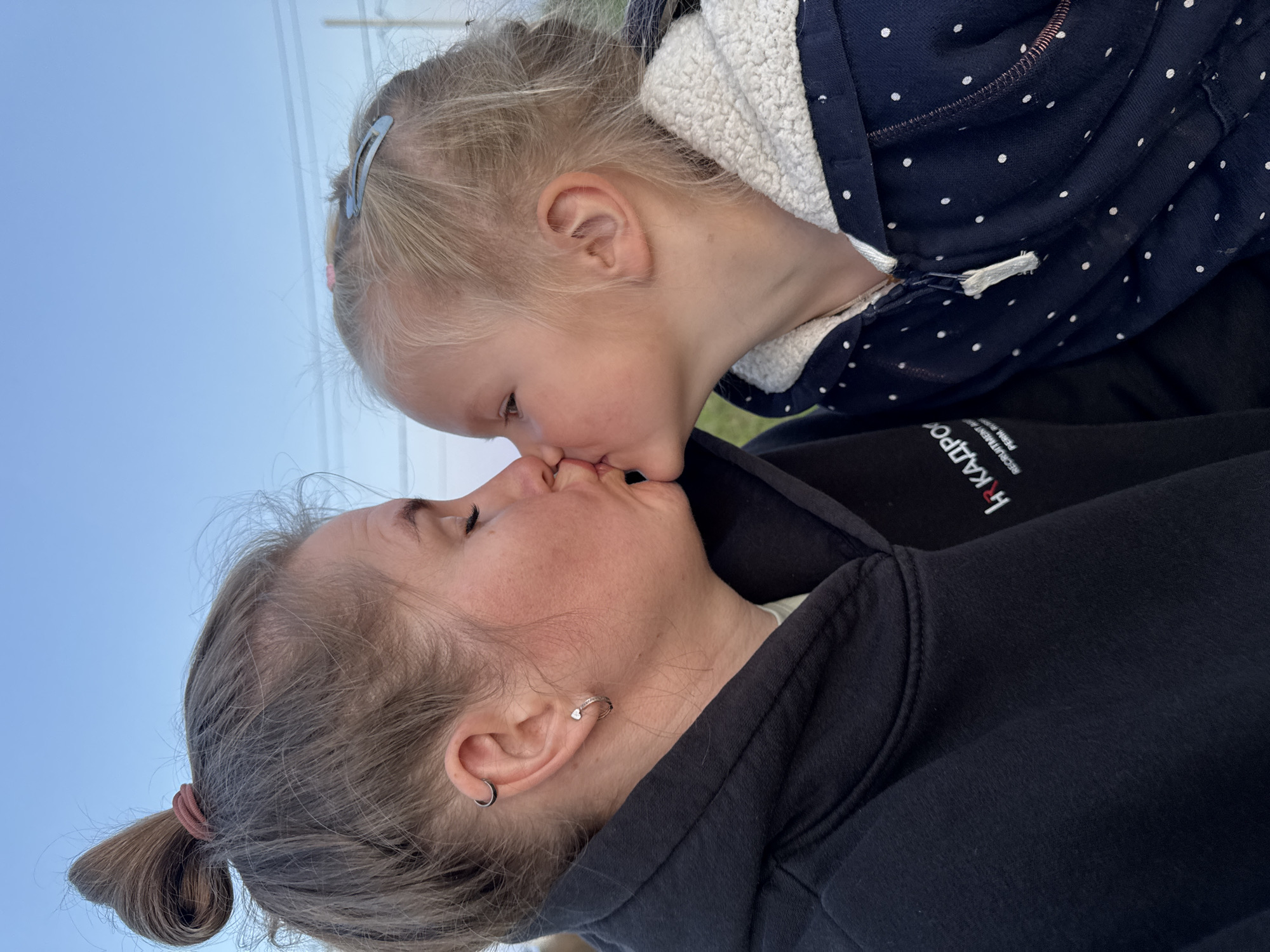 A close-up profile shot shows two people, an adult and a child, sharing a kiss against a clear blue sky. The adult, on the left, has their eyes closed and is wearing a black hooded sweatshirt. A small heart-shaped earring is visible in their left ear. The child, on the right, is also closing their eyes as they lean in for the kiss. They have blonde hair with a blue clip and are wearing a navy blue hooded sweatshirt with white polka dots. The background suggests an outdoor setting, possibly a field or open area, with a hint of distant structures or utility poles visible at the very top of the frame. The image is shot from a slightly low angle, with the focus on the faces and their tender interaction. The location is identified as Ферма, Russia.