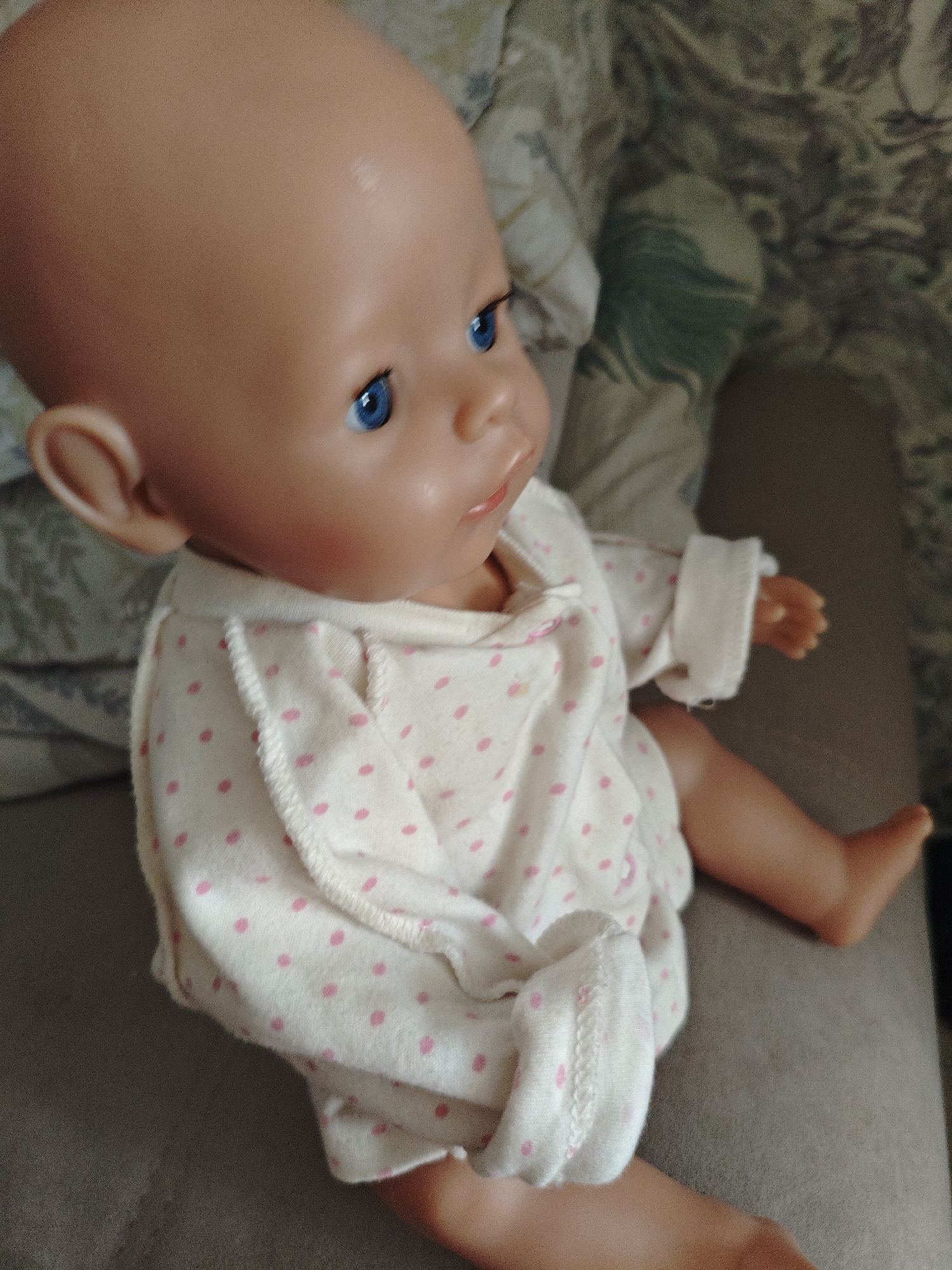 A baby doll with blue eyes and a bald head wears a white outfit with pink polka dots. The doll is lying on its side on a gray surface, with its right arm extended. In the background, a patterned fabric is visible. The image appears to be taken indoors. There is no information available about the location in the city of Berdsk municipality, Russia.