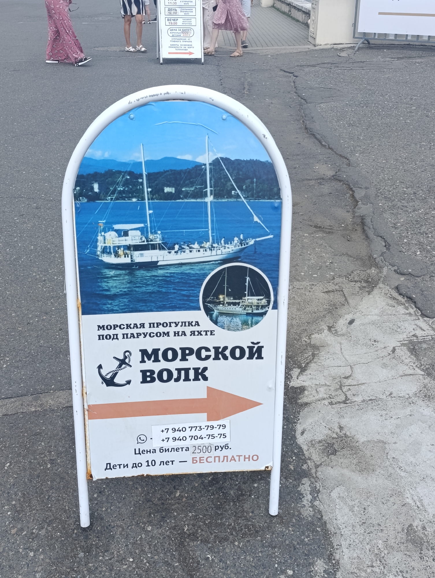 The image shows a weathered A-frame sign standing on an asphalt surface. The sign is advertising "MORSKOY VOLK" (Sea Wolf) boat tours. The sign features a large image of a sailboat on blue water and smaller images of boats. Text on the sign includes "MORSKAYA PROGULKA POD PARUSOM NA YAKHTE" (Sea trip on a yacht under sail), "MORSKOY VOLK" (Sea Wolf), phone numbers, "TSENA BILETA 2500 RUB." (Ticket price 2500 rubles), and "DETI DO 10 LET - BESPLATNO" (Children up to 10 years old - free). In the background, several people are walking on a paved area. The overall scene suggests a tourist location, likely a promenade or near a harbor in Sokhumi, Abkhazia.