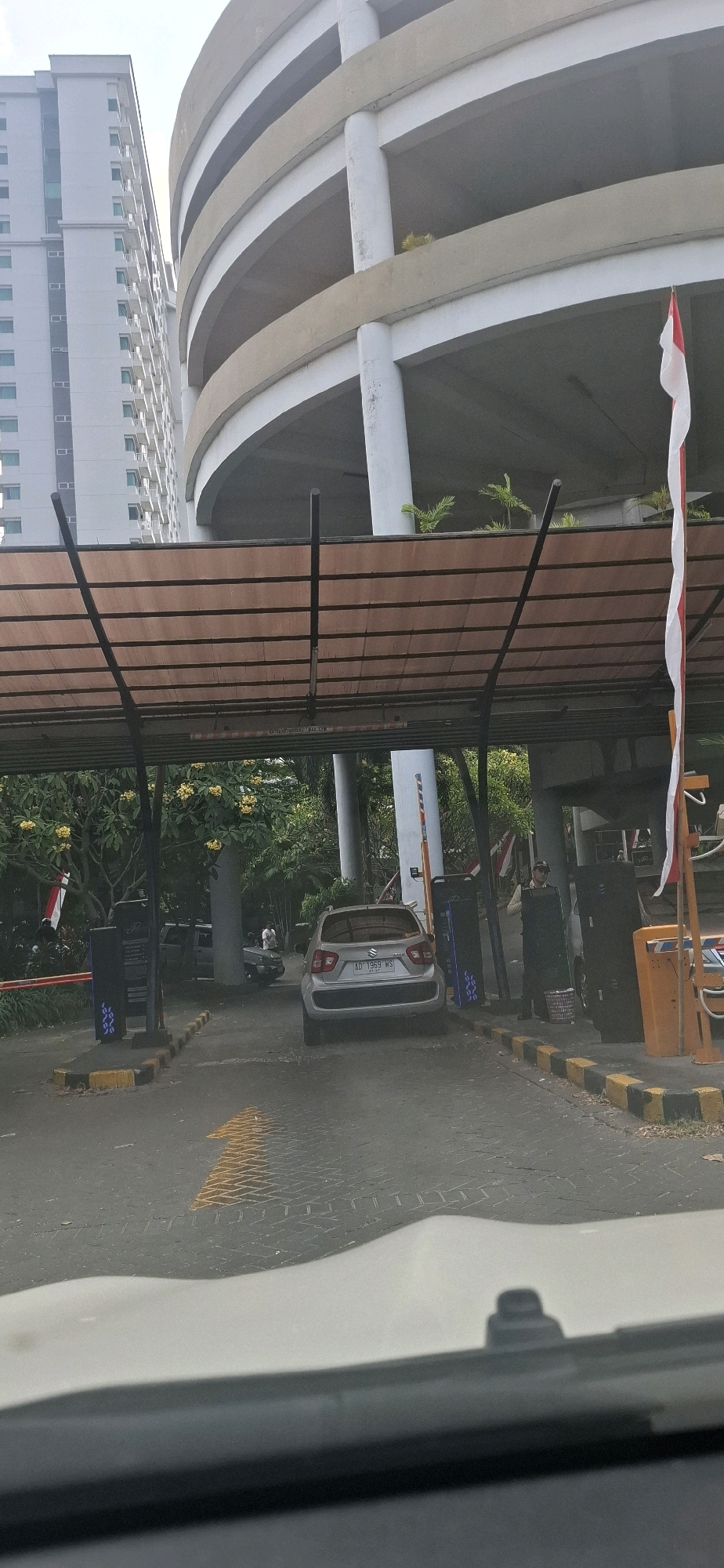 The image captures the entrance to a multi-level parking structure in Surakarta, Indonesia. A silver Suzuki Ignis is stopped at a gate, likely a payment booth. Behind the car, a person wearing a light-colored uniform and a cap stands near a kiosk. Red and white Indonesian flags are visible, suggesting a national holiday or event. The parking structure itself is a prominent feature, with its distinctive curved design and concrete pillars. A tall, modern building stands to the left of the parking structure, indicating an urban setting. The ground is paved with interlocking bricks, and yellow and black striped bollards line the entrance. The overall scene suggests a busy, functional urban environment with a focus on vehicle access and management.