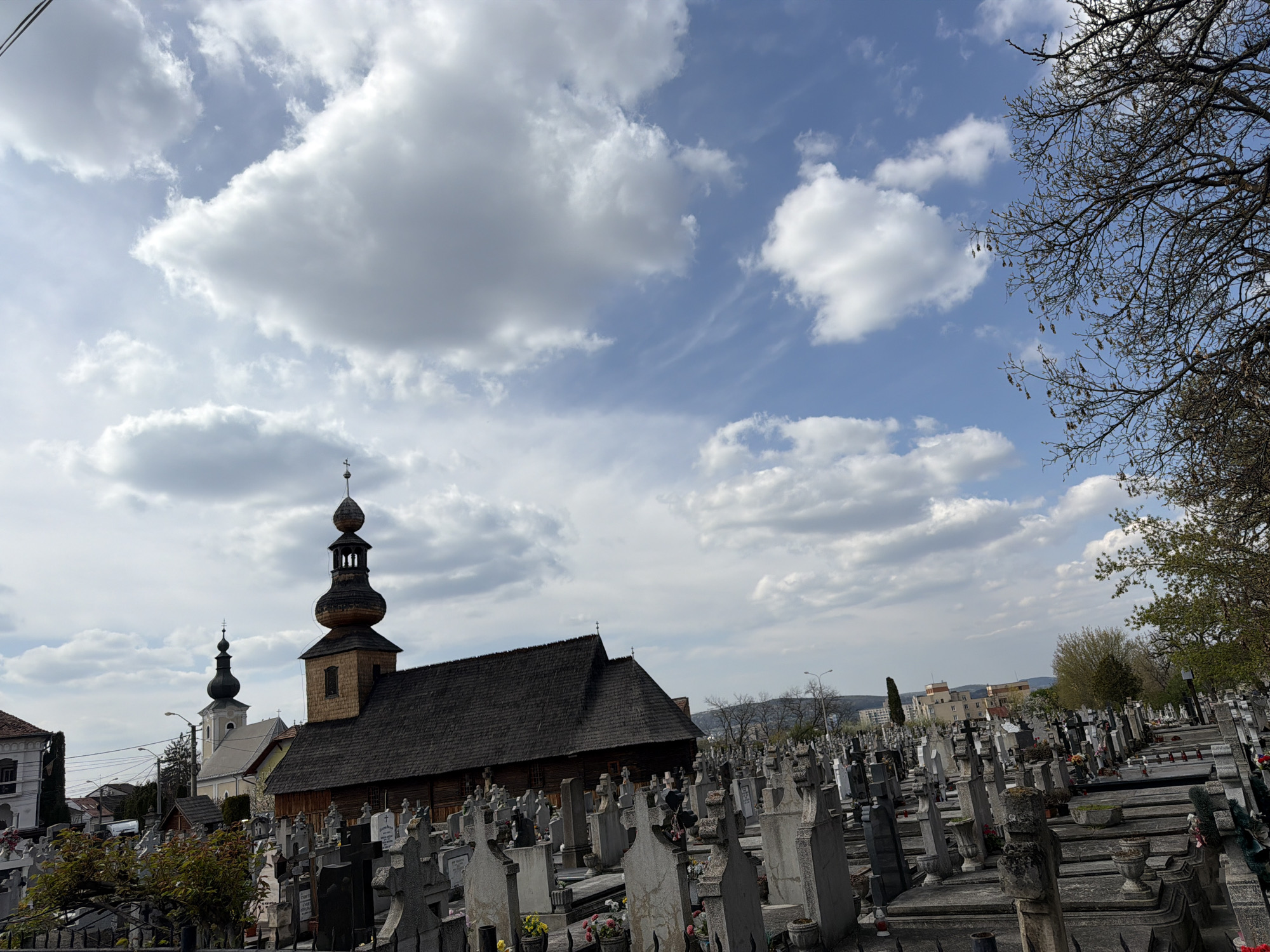 The image depicts a cemetery in Târgu Mureș, Romania, with a wooden church prominently featured. The sky is partially cloudy with a mix of white clouds and blue sky. The cemetery contains numerous tombstones and gravesites, indicating a well-established burial ground. The wooden church has a dark roof and a tall steeple. Other buildings can be seen in the background, suggesting an urban setting. Some trees with bare branches are also visible. There are no people depicted in the image.