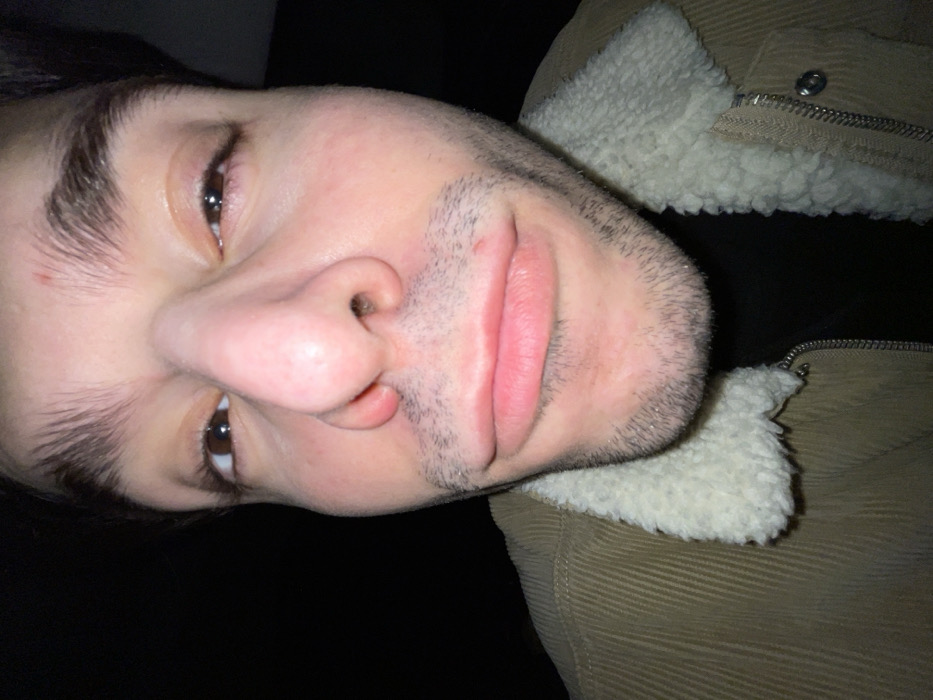 A close-up, angled shot of a person's face. The person has dark eyebrows, brown eyes, a prominent nose, and stubble on their chin and upper lip. Their lips are pink. Part of a tan corduroy jacket with a white, fluffy collar is visible on the right side. The background is dark, and the image is slightly tilted.