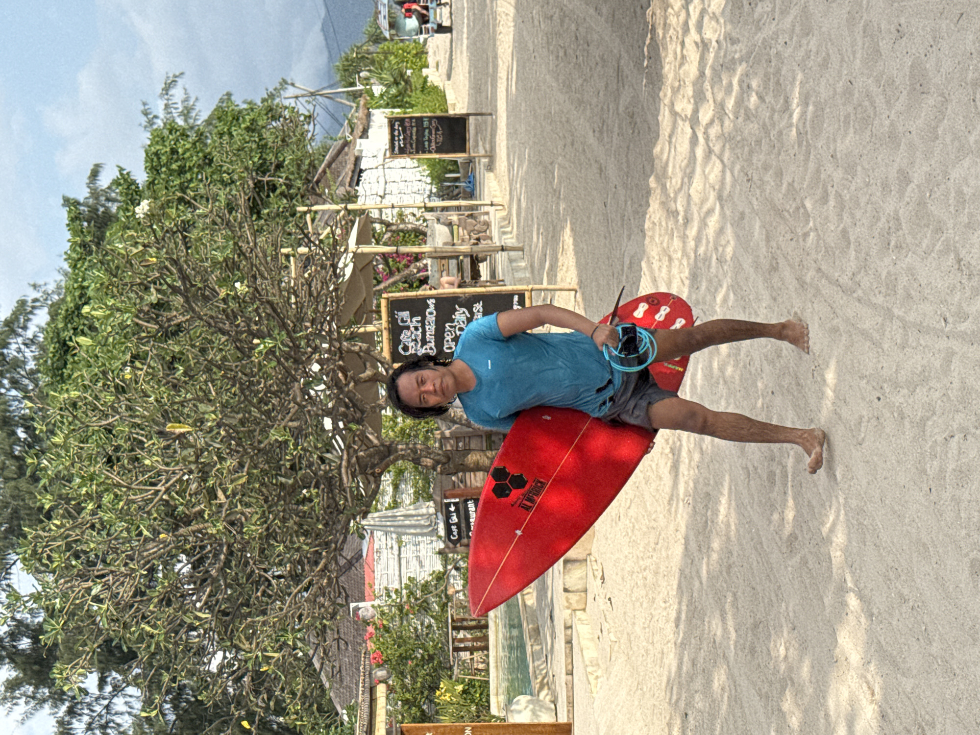 The image shows a man holding a red surfboard on a sandy beach. The surfboard has the logo "AL MERRICK" with some other logo. The man is wearing a light blue shirt and gray shorts. Behind him, there is a tree, and behind the tree, there are buildings and signs written "Cafe Gi Beach Bungalows open Daily st", "Cape Gi aurant", "Love Toyike 115k. The scene is outdoors, and the overall impression is that it is a sunny day at a beach resort. The man is standing in front of some structures that have signs on them. The location is city of Unknown, Indonesia.